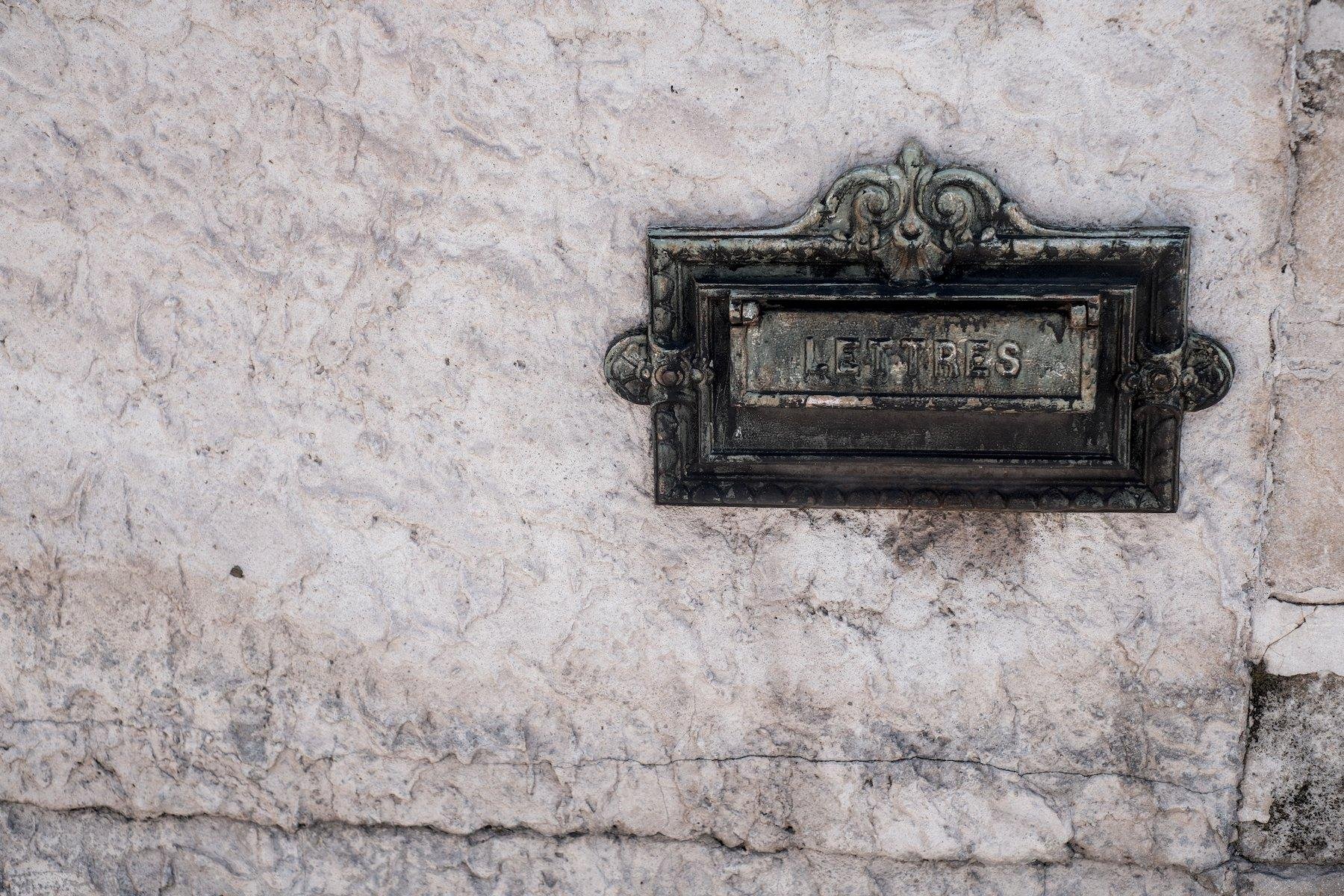 Horizontal photo. In a wall is embedded the opening of a metallic letterbox, which looks ancient and has fancy curly decoration on all the sides with an extra one on the upper side. The flap is engraved with ”LETTRES”