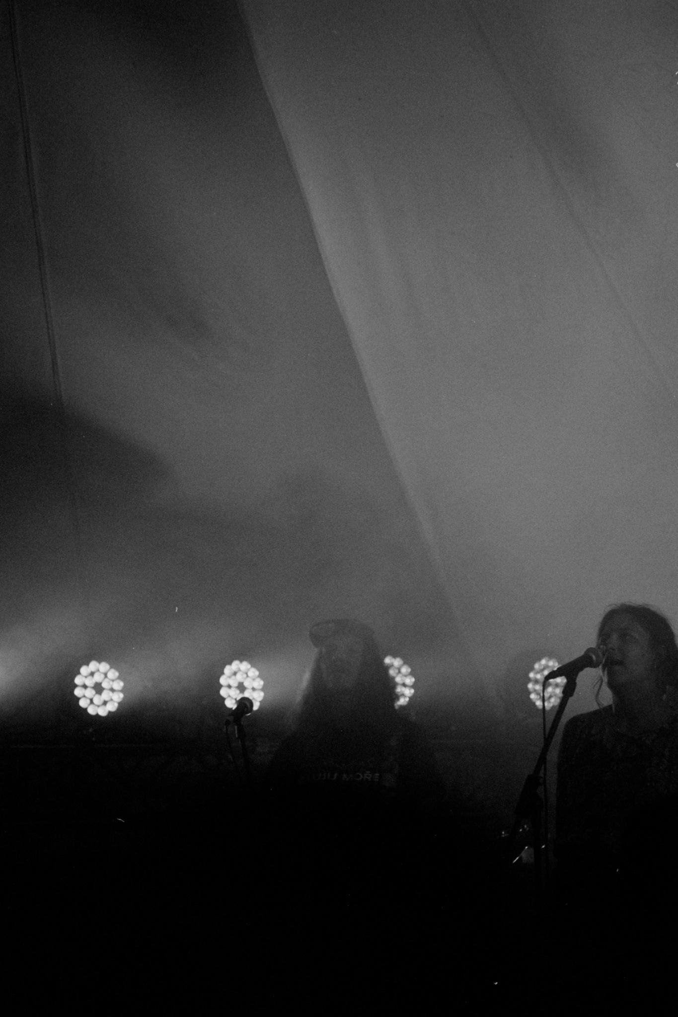 Black-and-white concert photo with two singers at microphones near the bottom right, partially silhouetted by stage haze. Four circular stage lights glow in a row behind them, with a large draped backdrop filling most of the frame.