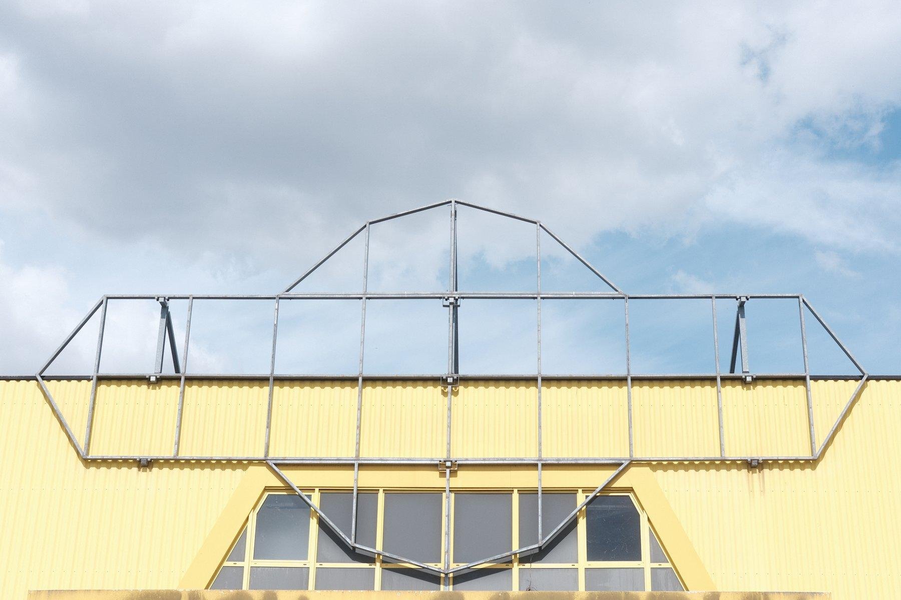 Photo horizontale. Sommet d’un mur jaune pâle, avec l’armature d’une enseigne qui a été démontée. On reconnait la forme du logo de Bricorama, sous un ciel nuageux avec un bout de bleu