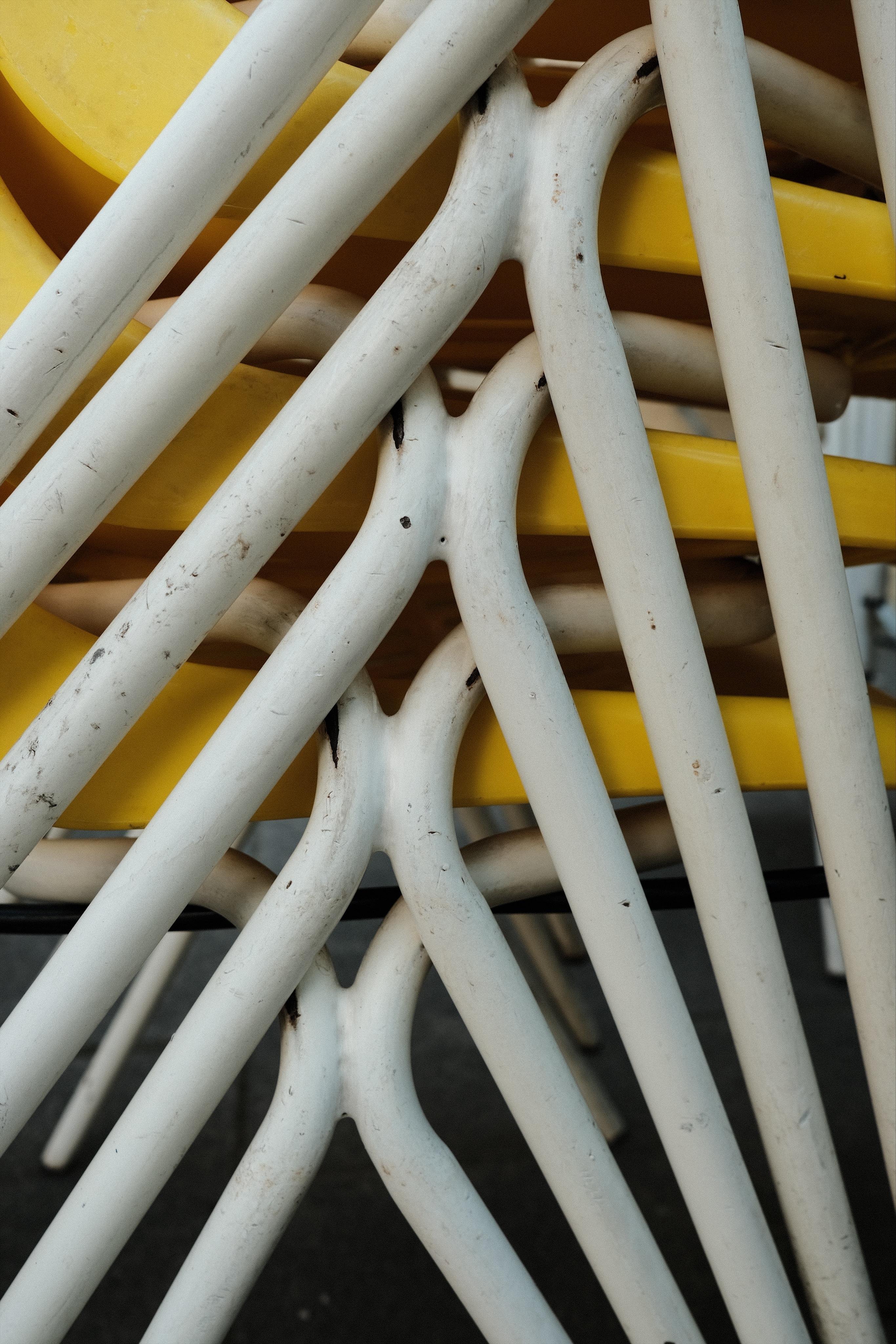 A stack of chairs with yellow plastic seats and white metal frame/legs. This is a detail shot of the white metal frames of several stacked chairs that create a complex mesh-like pattern. Between the white pipes parts of the yellow seats are visible.