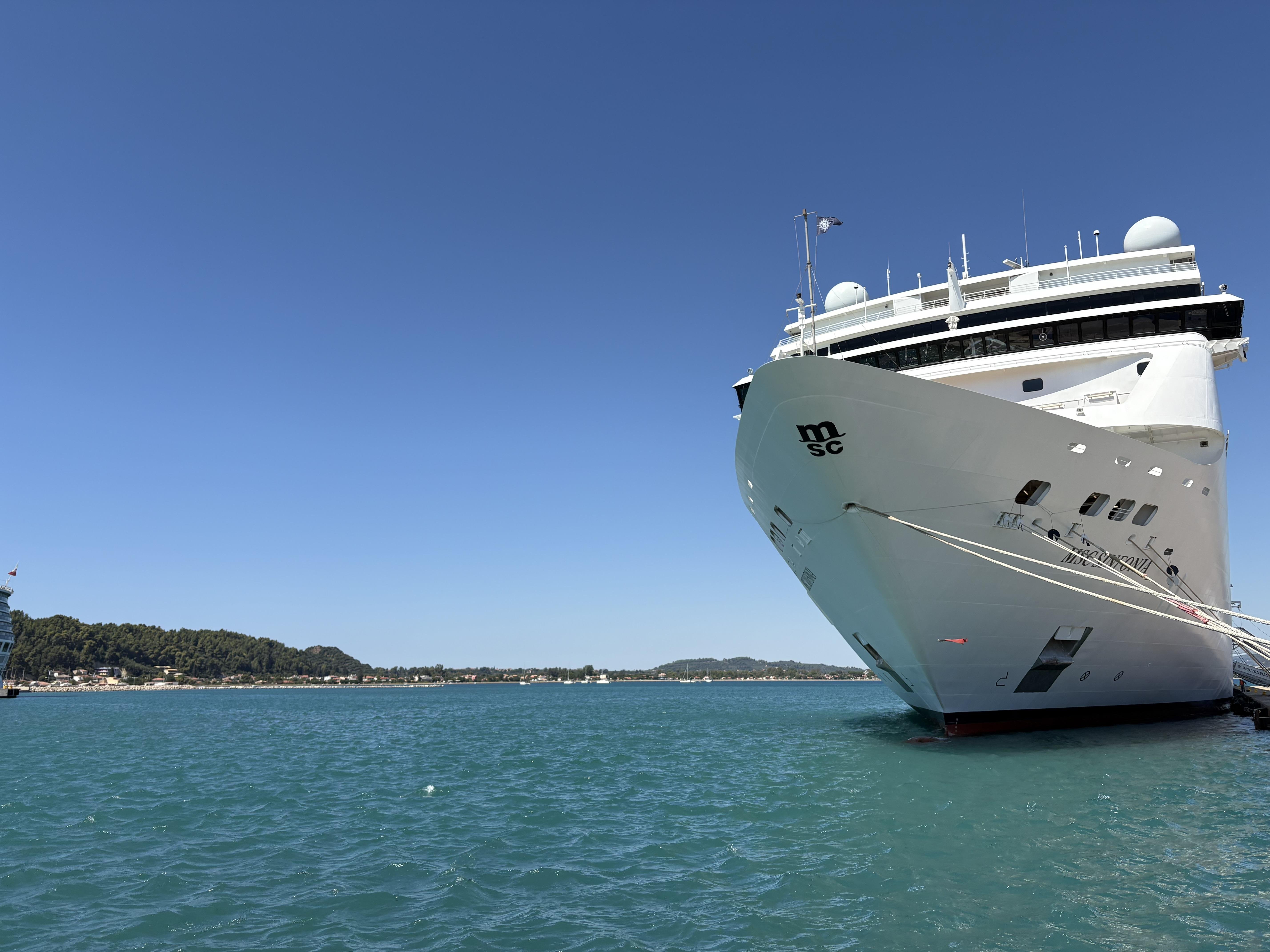 Large white cruise ship docked in a harbor, with clear blue sky and calm water. The bow features the letters "MSC". A tree-lined coastline is visible on the left, extending into the horizon.