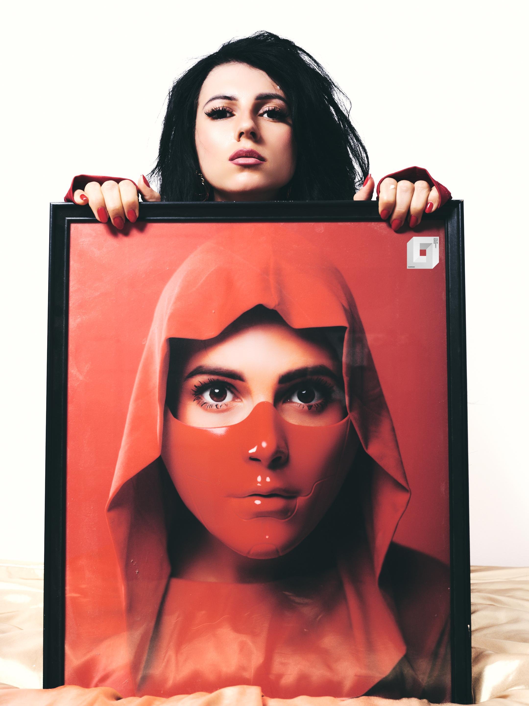 Woman with dark hair holds the top edge of a large black-framed portrait; the portrait shows a close-up of a face wearing a red hood and red mask against a red background, set on a light surface with a white backdrop.