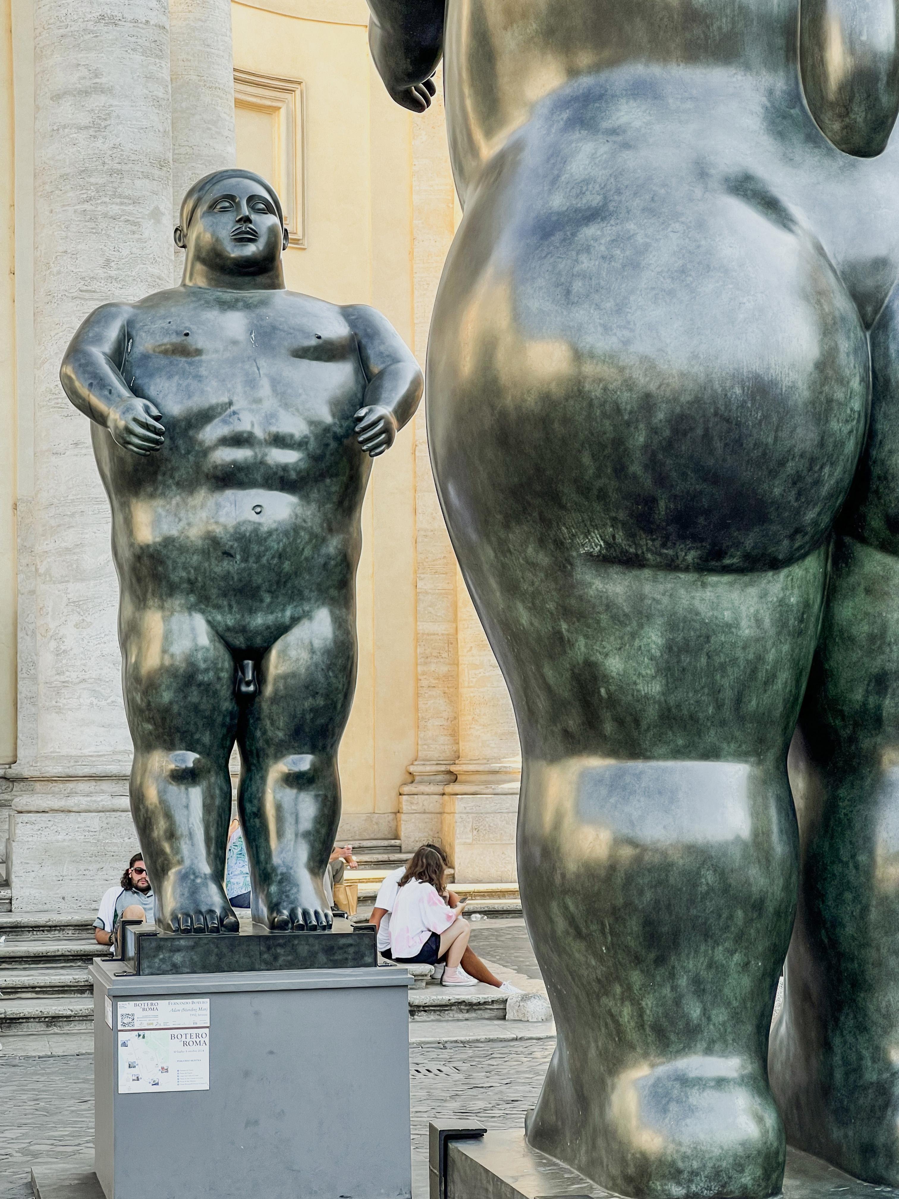Grandi sculture di figure umane, caratteristiche dello stile di Fernando Botero, noto per raffigurare persone e figure con volumi esagerati. Le sculture sono di colore scuro e sono situate in un'area esterna con uno sfondo di elementi architettonici. Ci sono anche persone sedute o che si riposano sullo sfondo.
