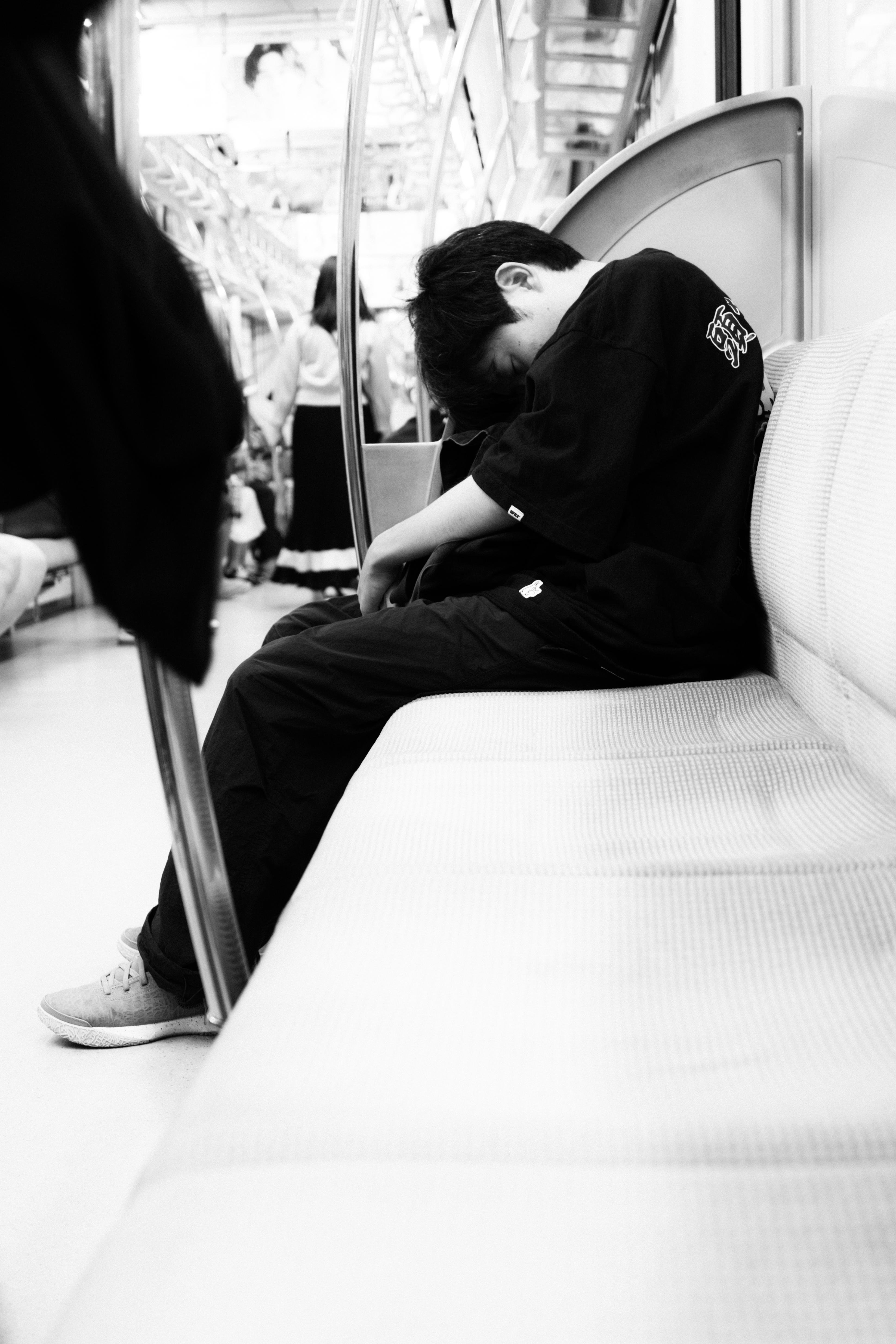 Young person in dark clothing sleeping with their head down on a subway seat. The subway is moderately crowded with people standing in the background. The image is in black and white, highlighting the contrast between the person's clothing and the light-colored seat. A metal pole separates the viewer's perspective from the rest of the carriage.