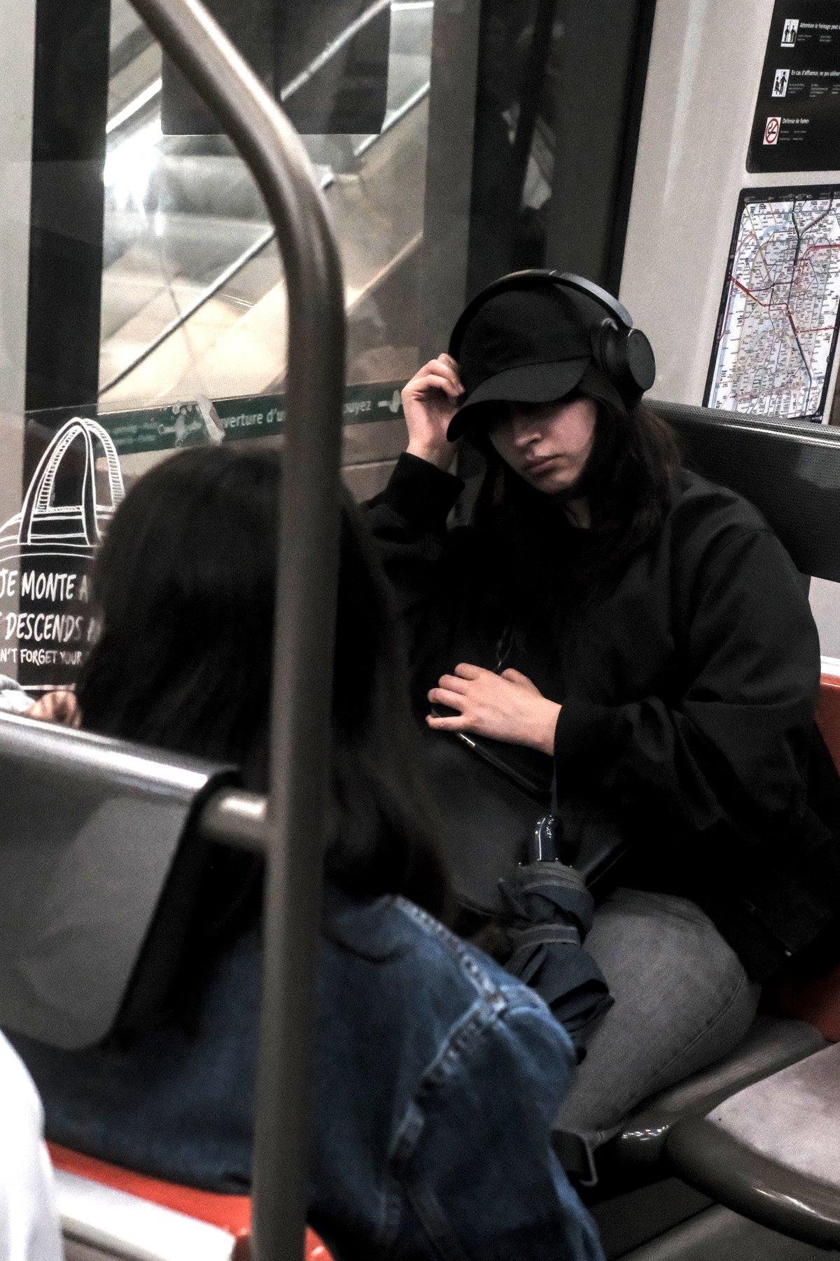 Photo verticale. Dans un carré de 4 sièges du métro, une jeune femme aux cheveux noirs sous une casquette noire, sous un casque audio noir, avec un jean noir, a les yeux cachés par la visière, la main gauche aggripant son téléphone plaqué contre son sac, la main gauche réajustant le casque