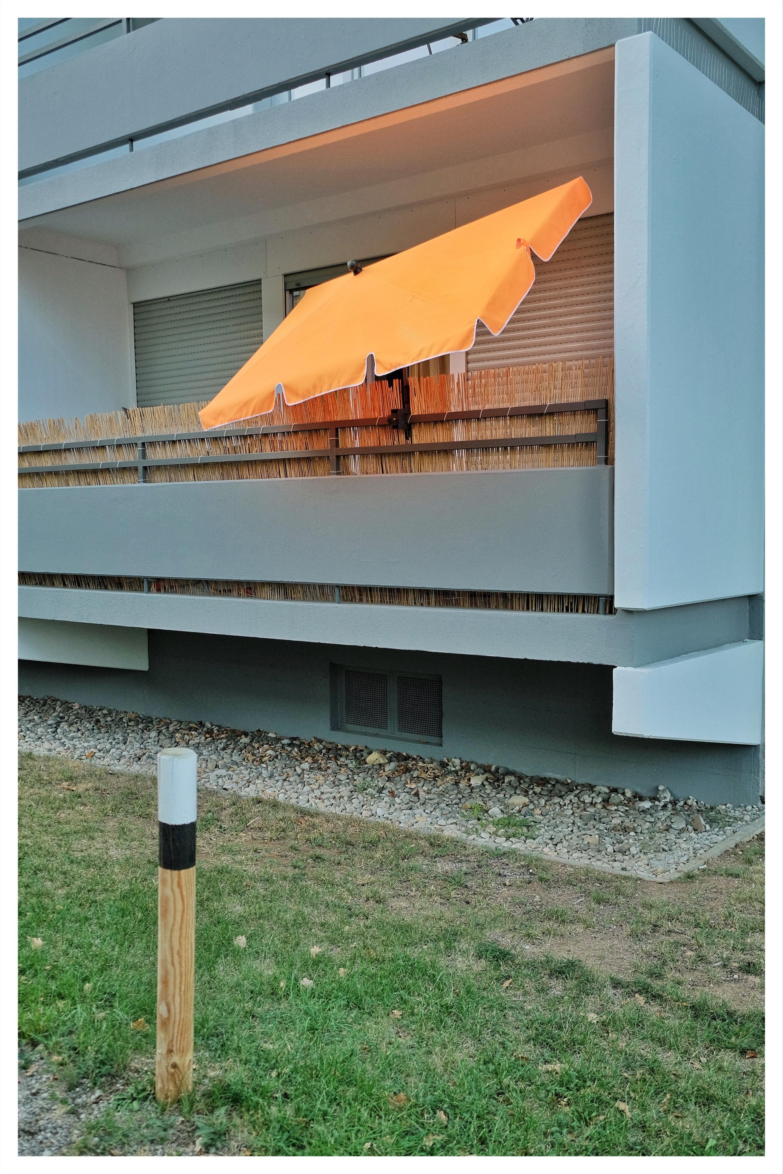 Orange patio umbrella leaning over a gray apartment balcony with bamboo screening and closed shutters; grassy yard and a small wooden post in the foreground.