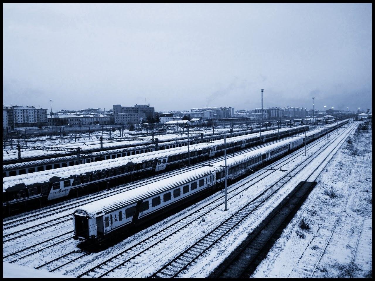 This image depicts a railway yard covered in snow, with several train tracks and trains visible. The landscape appears wintry, with buildings in the background under a cloudy sky. The scene conveys a cold and quiet atmosphere typical of a snowy day.
