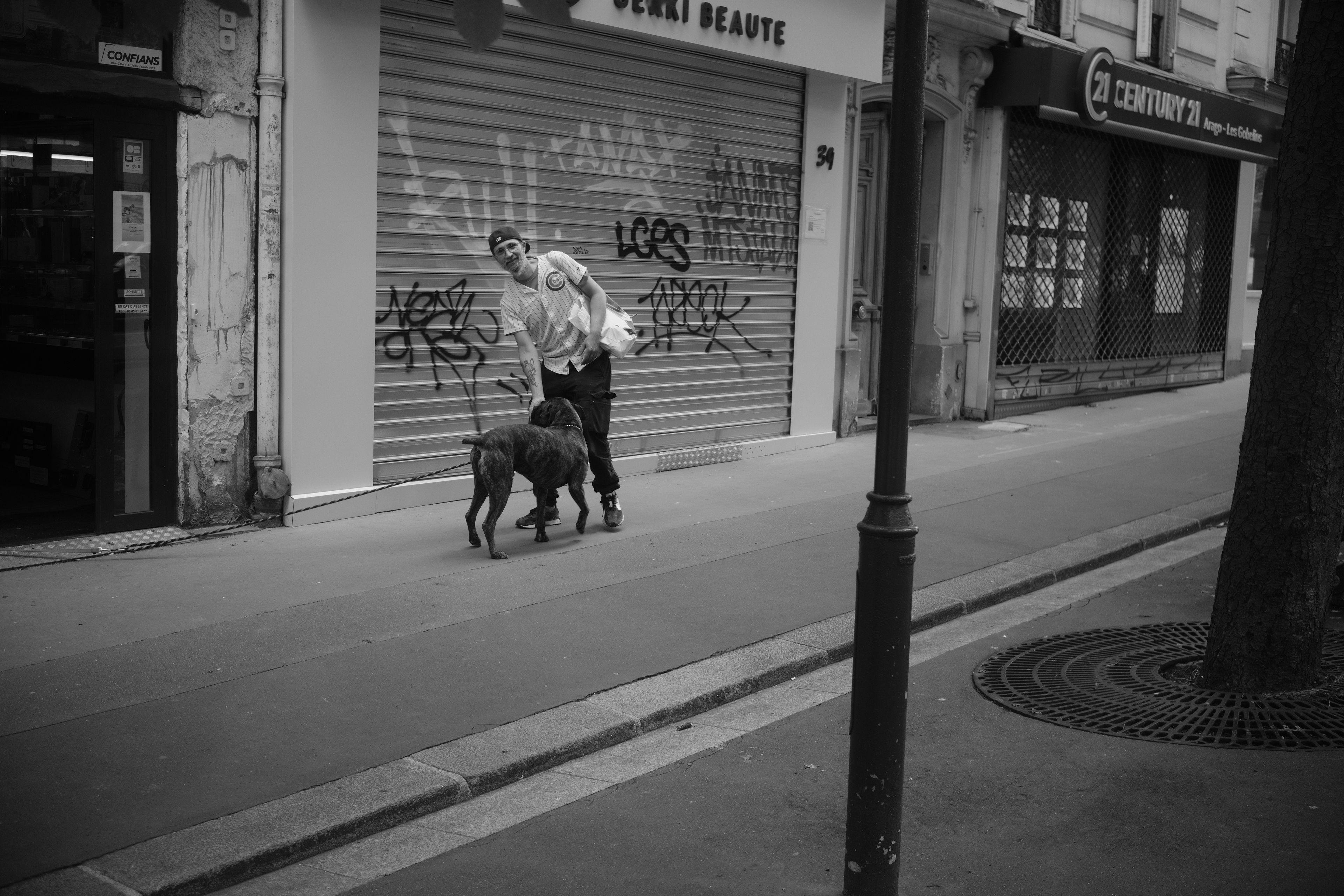 Photo noir et blanc dans la rue, un grand chien noir va à la rencontre de son maître qui le caresse en regardant la caméra. Il a une casquette portée à l’envers, une chemise à rayures, un pantalon noir et des baskettes