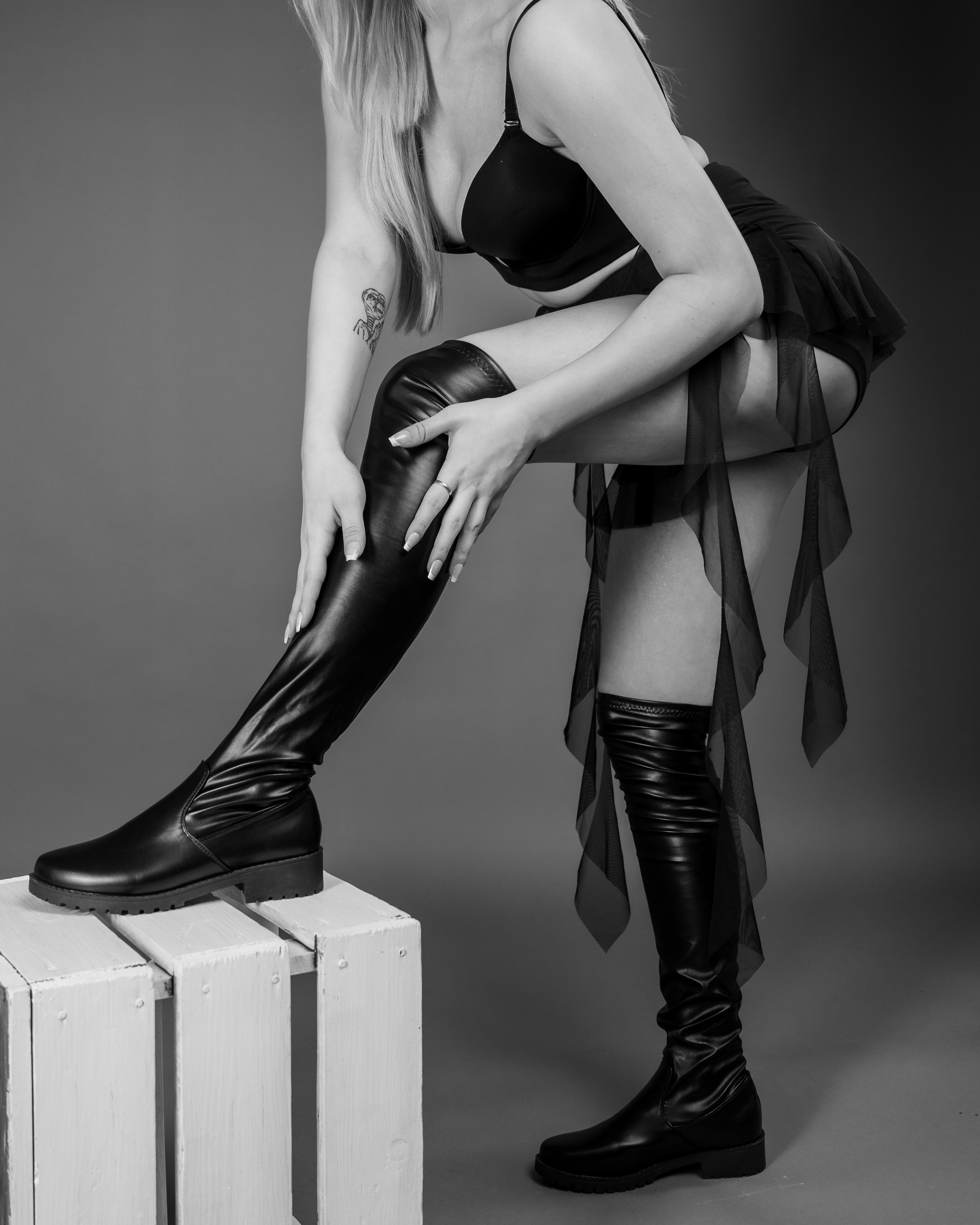 Woman wearing a black bra, skirt, and thigh-high leather boots, with long hair cascading over her shoulder. She is bent forward, adjusting her boots on a white wooden crate, with sheer fabric draping from her skirt. Tattoo visible on her left arm.