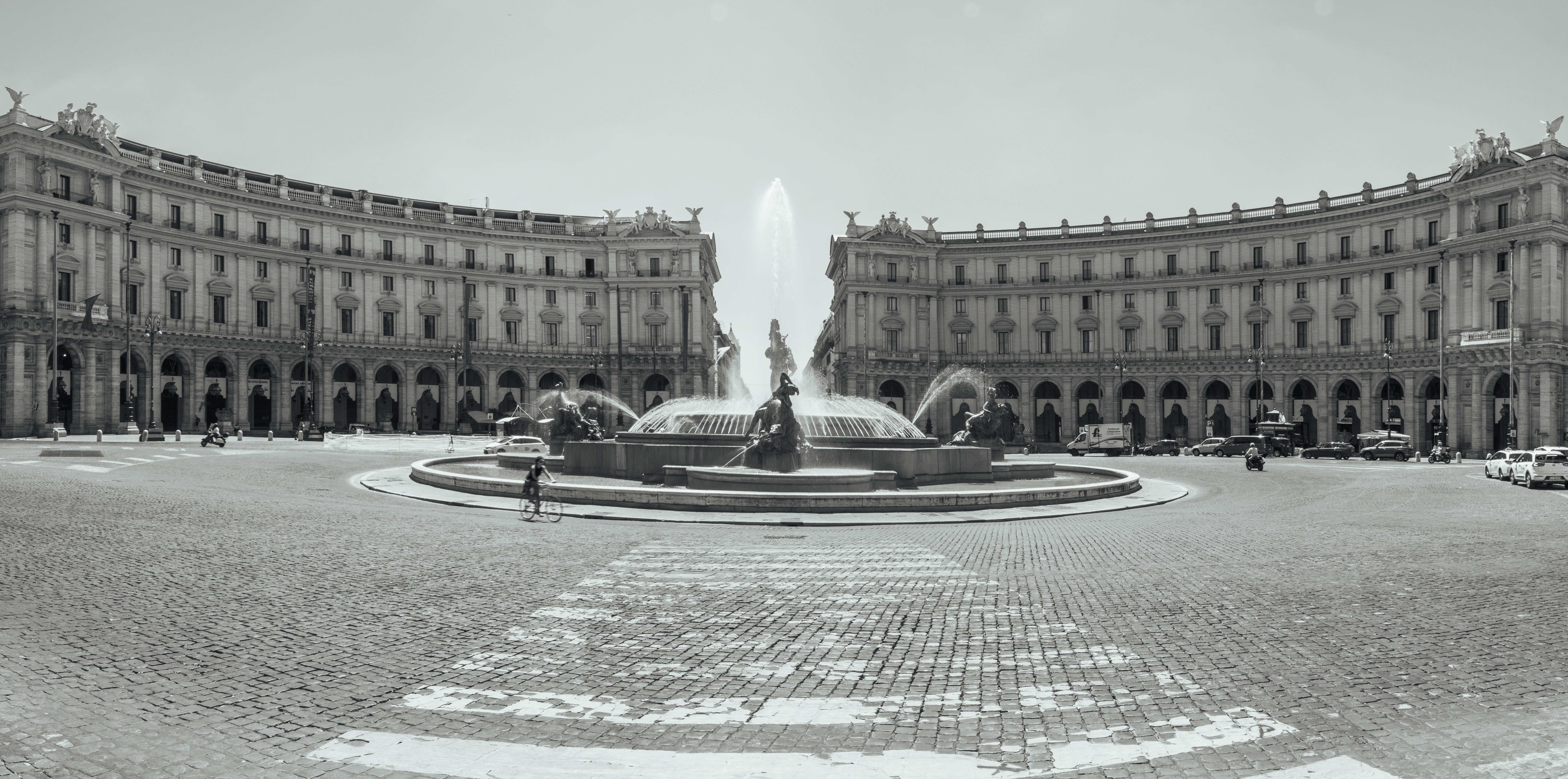 L'immagine mostra Piazza della Repubblica a Roma, Italia. Presenta una grande piazza circolare con una fontana centrale nota come la Fontana delle Naiadi. Gli edifici circostanti sono maestosi e architettonicamente impressionanti, contribuendo all'atmosfera storica e culturale del luogo. La scena sembra essere stata ripresa durante il giorno ed è presentata in bianco e nero.
