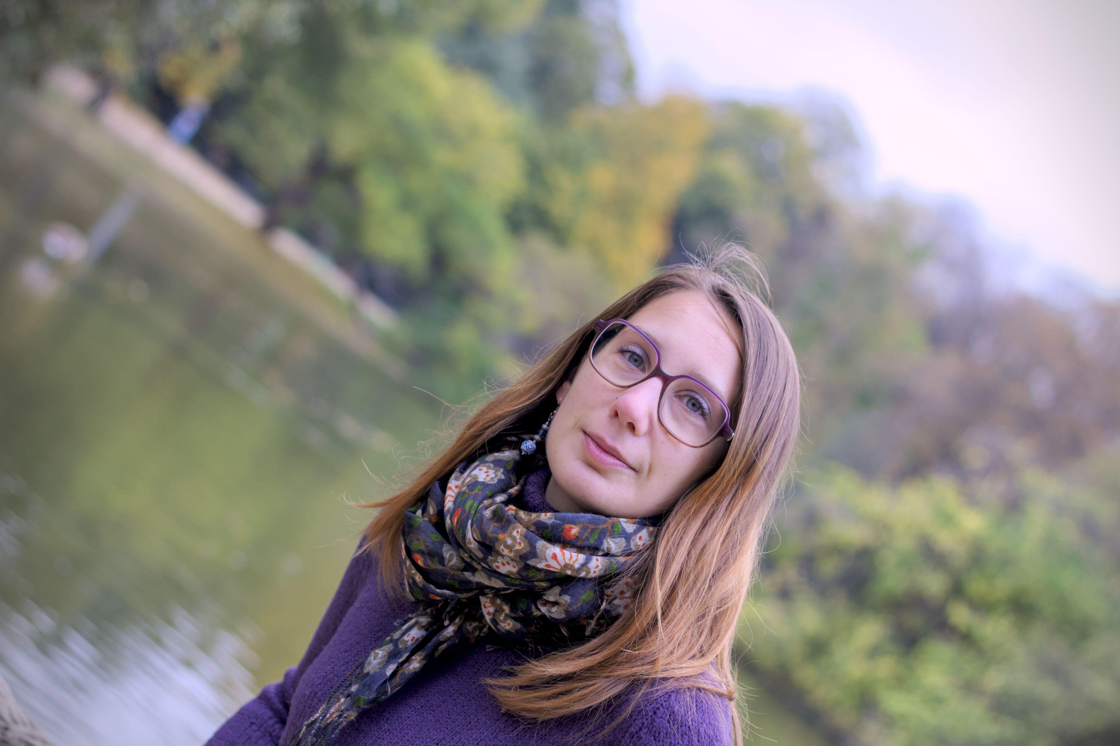 Photo format paysage, à 45⁰, montrant une jeune femme châtain portant des lunettes violettes, un pull violet et un foulard au motifs fleuris bleu sombre, blanc, vert et rouge. Elle regarde la caméra avec un léger sourire aux lèvres, sur fond de lac et arbres