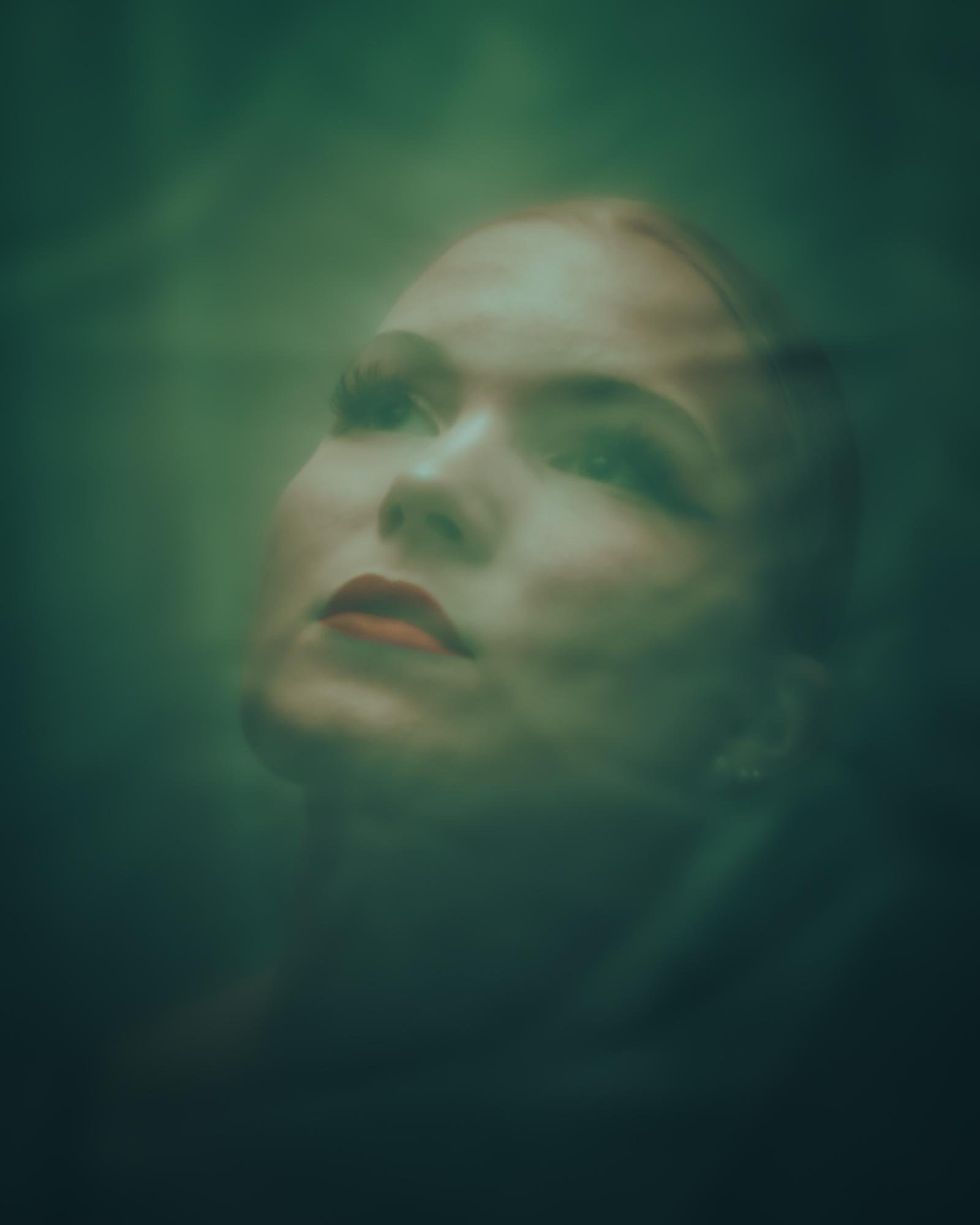 The image is an artistic photograph of a person's face, partially obscured and blurred, likely underwater or behind a translucent surface. The colors are predominantly greenish, creating a dreamy or ethereal effect.