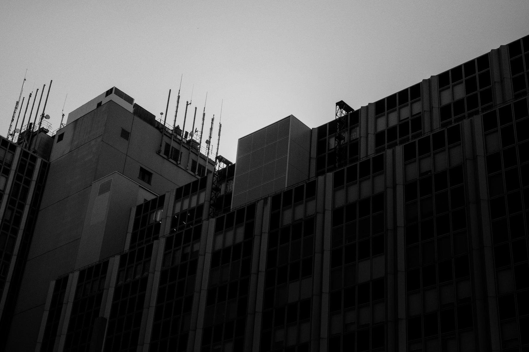 Photo horizontale en noir et blanc. Contre plongée sur un grand bâtiment qui cache le soleil dont on voit le halo derrière.