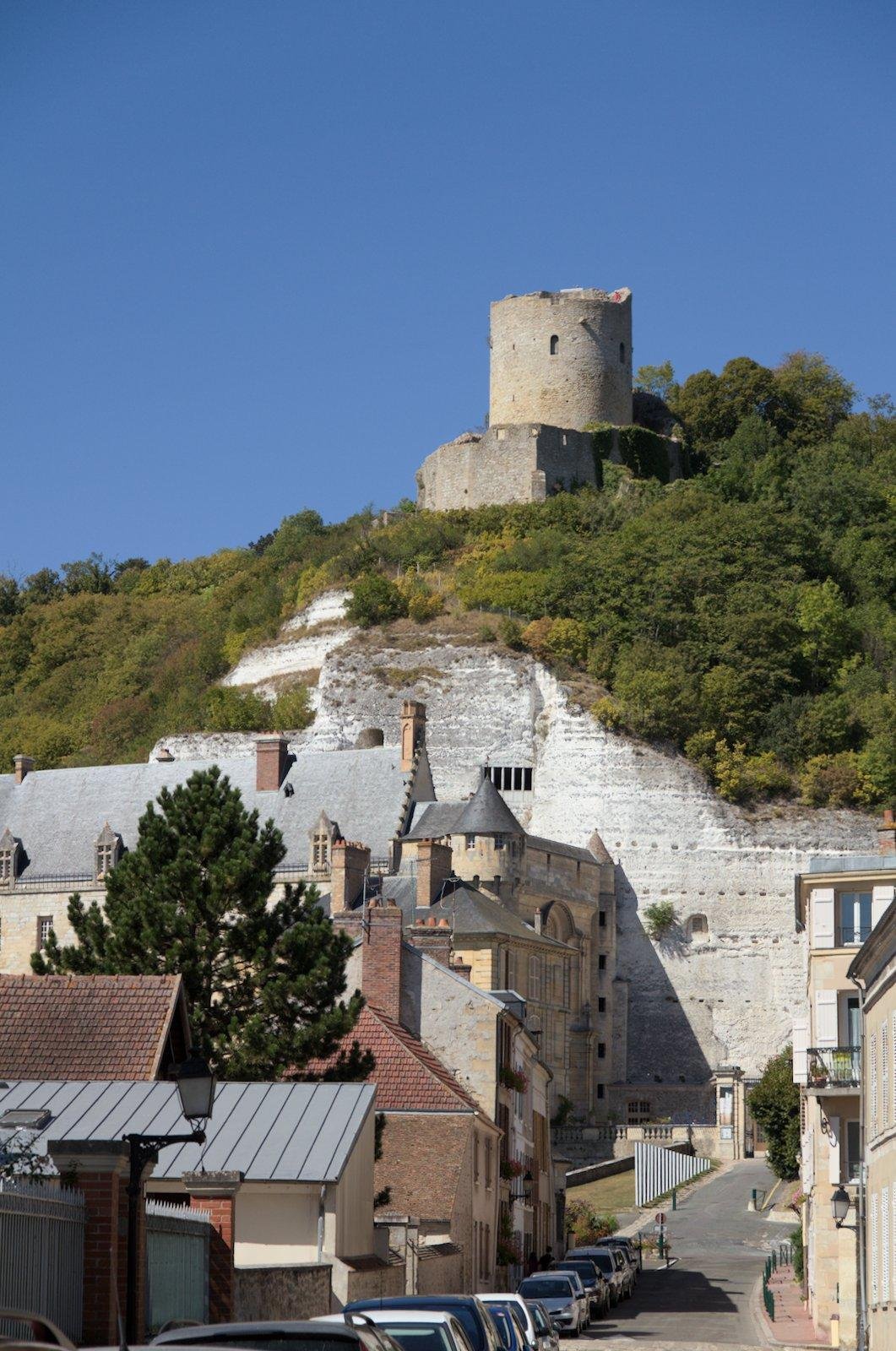 Photo format portrait montrant le donjon de la Roche-Guyon, en haut d’une falaise blanche avec plein de végétation dessus. En bas, une rue du village avec des maisons, le tout sous un ciel bleu
