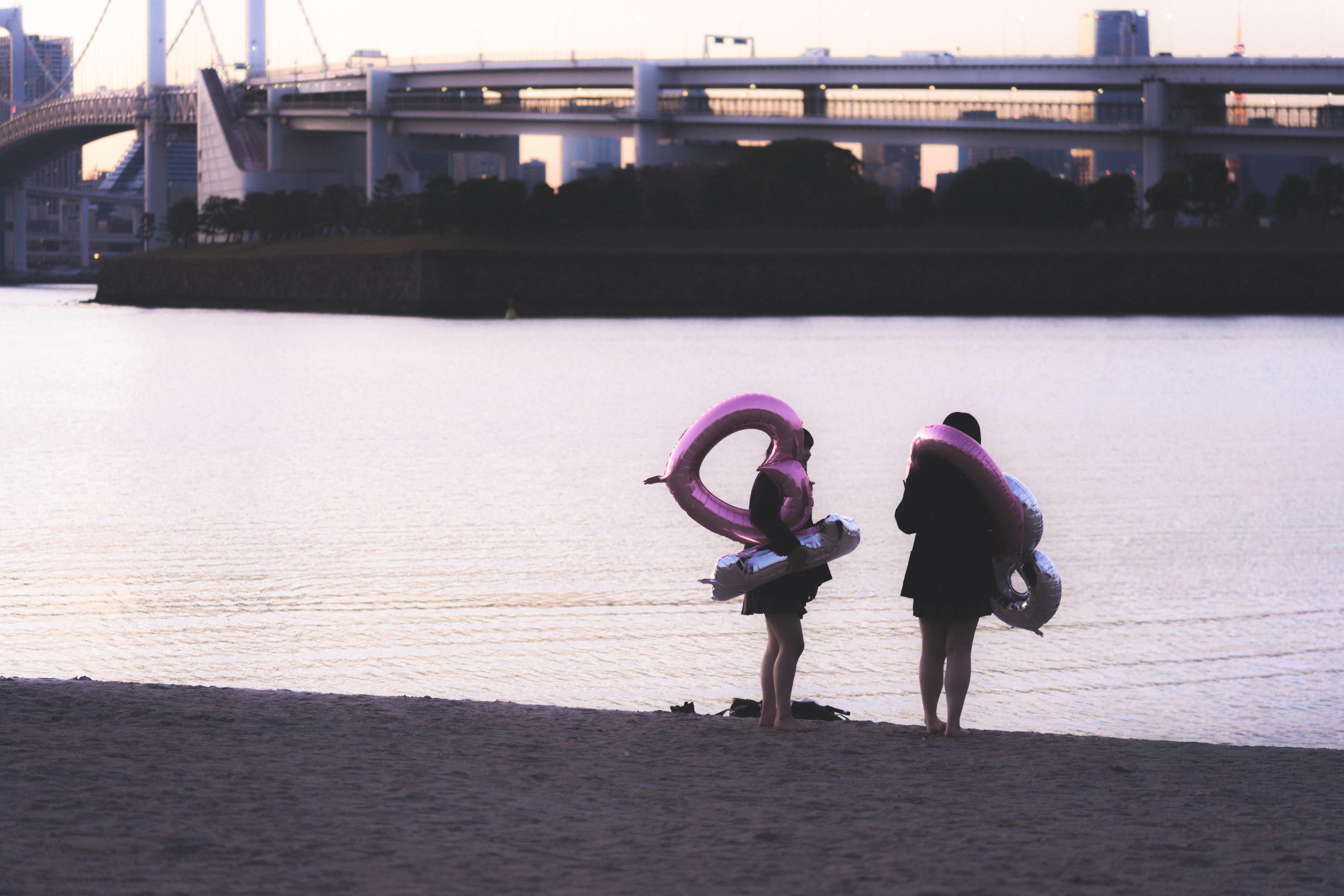 Two people stand on a sandy beach, each holding inflatable pink swim rings. A body of water stretches out in front of them, with a large bridge and skyline visible in the background at dusk. The scene is calm, with soft lighting suggesting early evening.