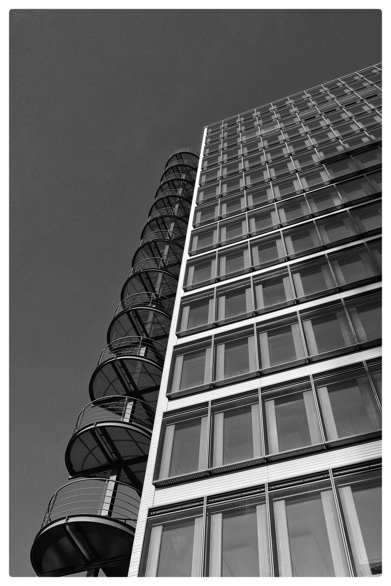 Black and white image of a modern building with a grid of rectangular windows on the right. An adjacent cylindrical staircase with a metal railing spirals upward on the left. The perspective is angled upward, emphasizing the height of the structures against a clear sky.