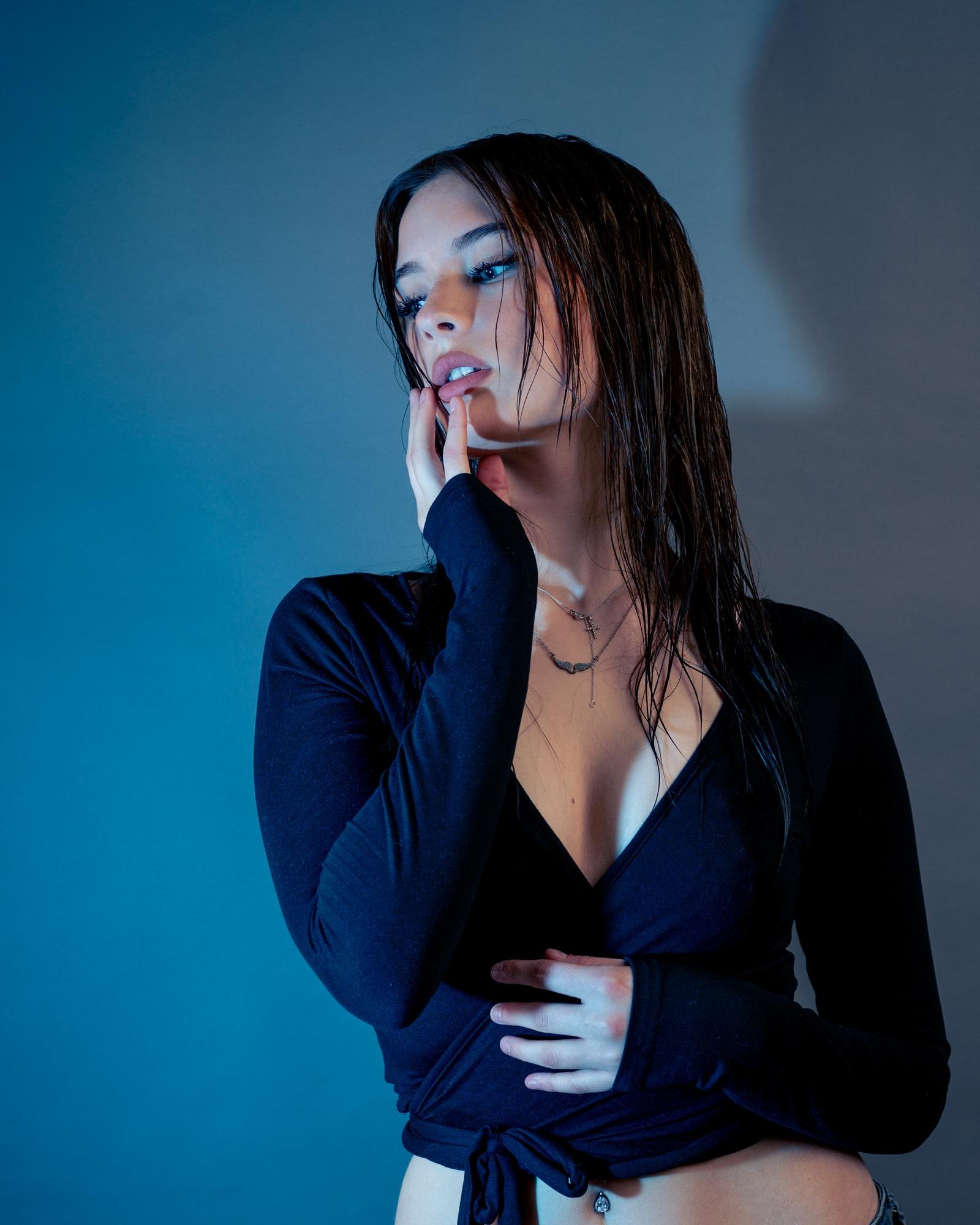 Woman with wet hair wearing a black top with long sleeves, posing with her right hand touching her chin. Her outfit includes a v-neck and a tie at the waist. Necklace displayed around her neck. Gradient blue background creating a soft shadow.