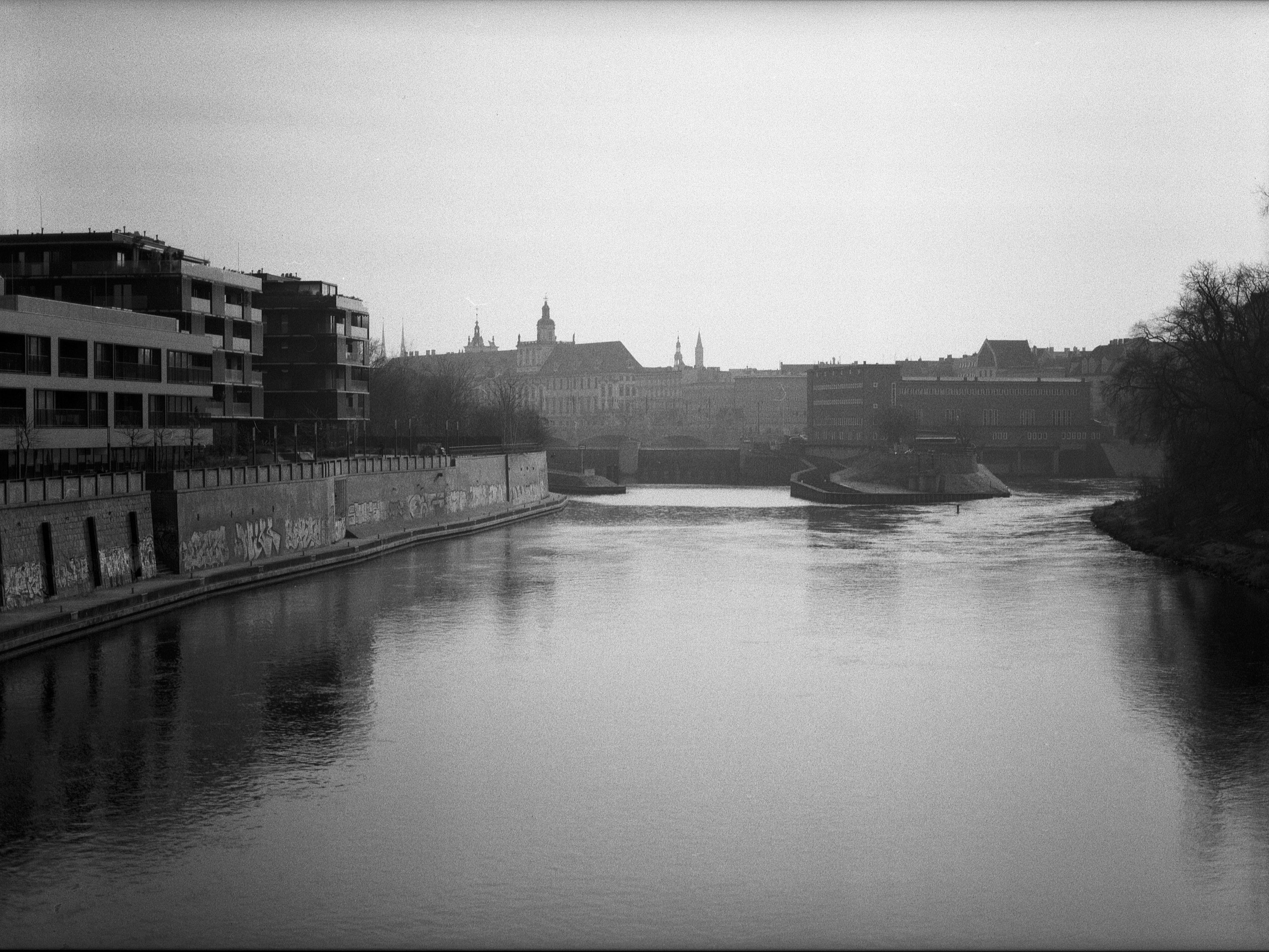 Black and white image of a river running through an urban area, flanked by modern buildings on the left with visible graffiti on the walls.