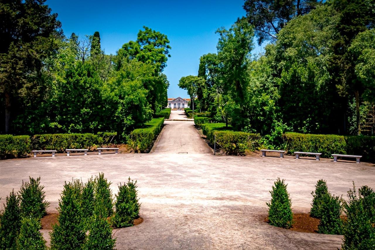 Wide, straight pathway lined with manicured bushes and benches, leading to a distant building with a red roof. Tall trees and lush greenery surround the path under a clear blue sky.