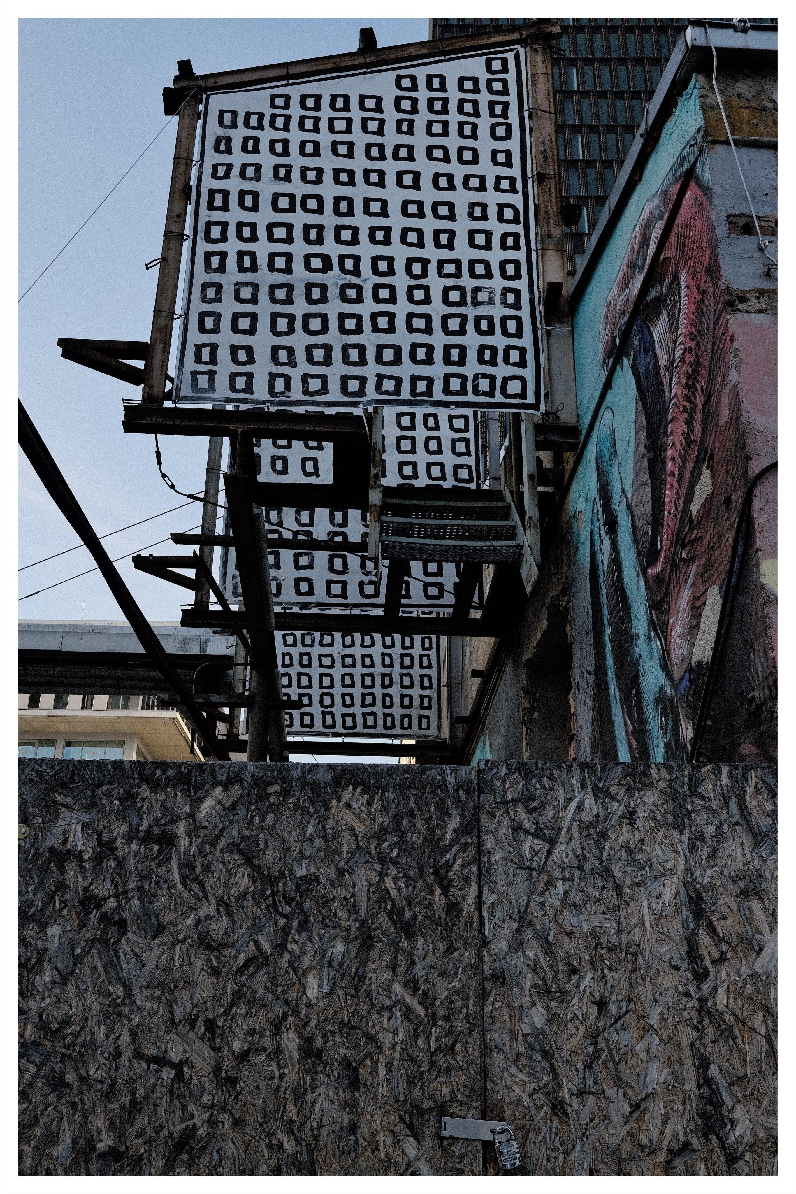 Stacked rectangular panels printed with rows of black square outlines hang from a metal scaffold beside a building with a colorful mural; plywood boarding spans the lower half, with sky and nearby buildings in the background.