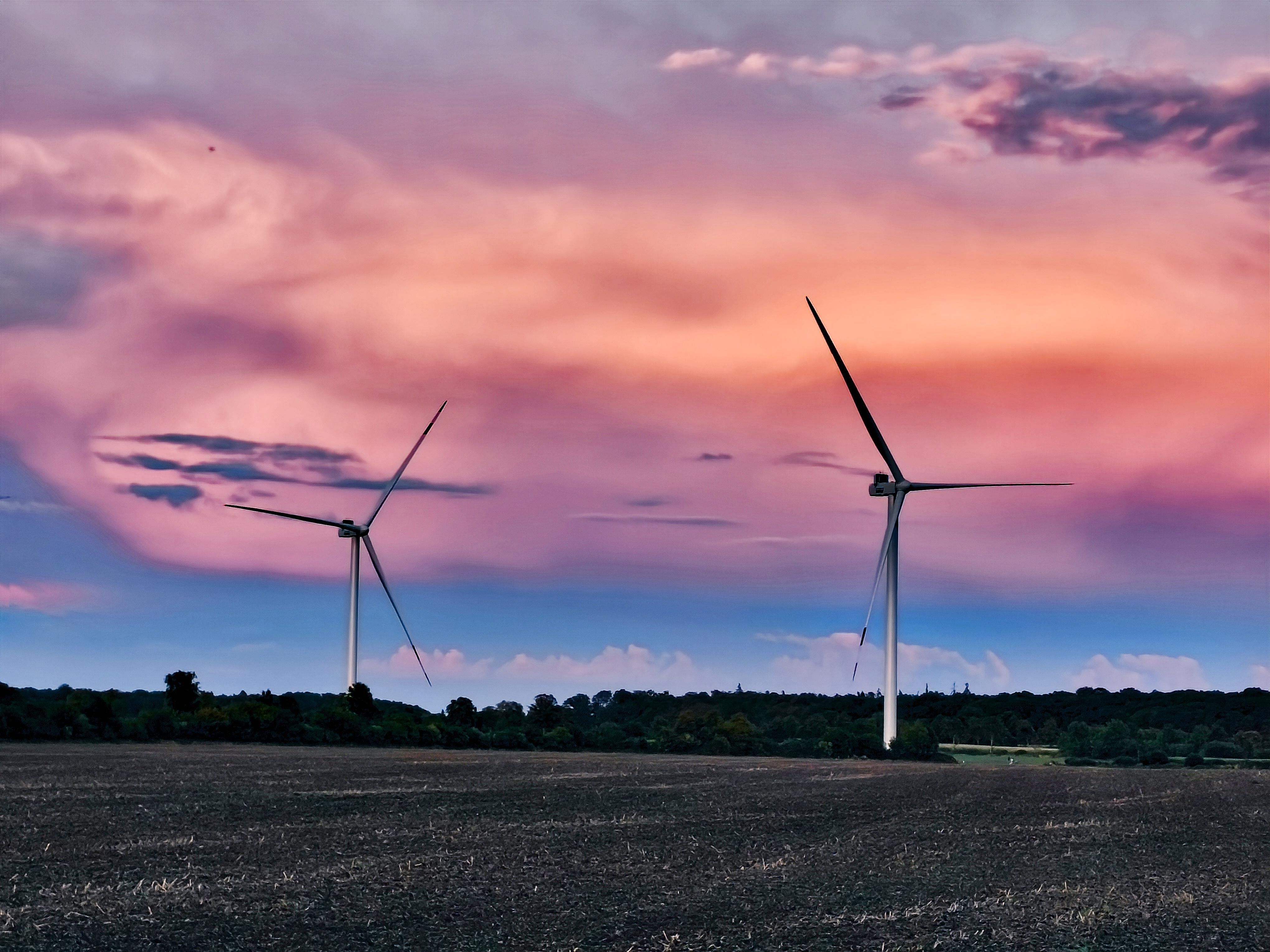 Two wind turbines in an open field with a dark tree line on the horizon, under a pink and purple cloud-filled sky near sunset. 
### 
Zwei Windkraftanlagen auf einem offenen Feld mit einer dunklen Baumgrenze am Horizont, unter einem rosa und violetten, mit Wolken gefüllten Himmel in der Nähe des Sonnenuntergangs.