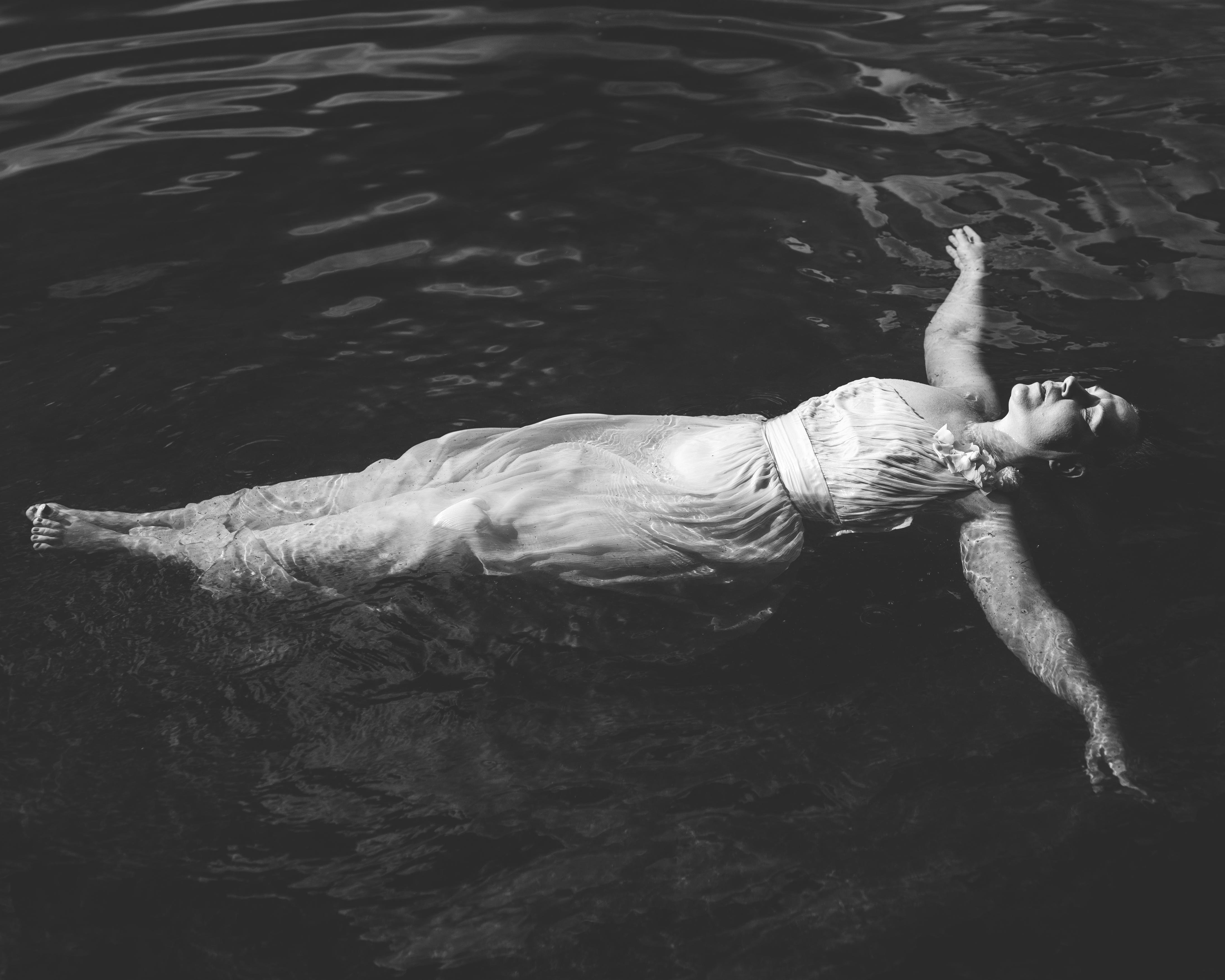The image depicts a person floating on water, wearing a dress. It is presented in black and white, giving it a serene and artistic feel.