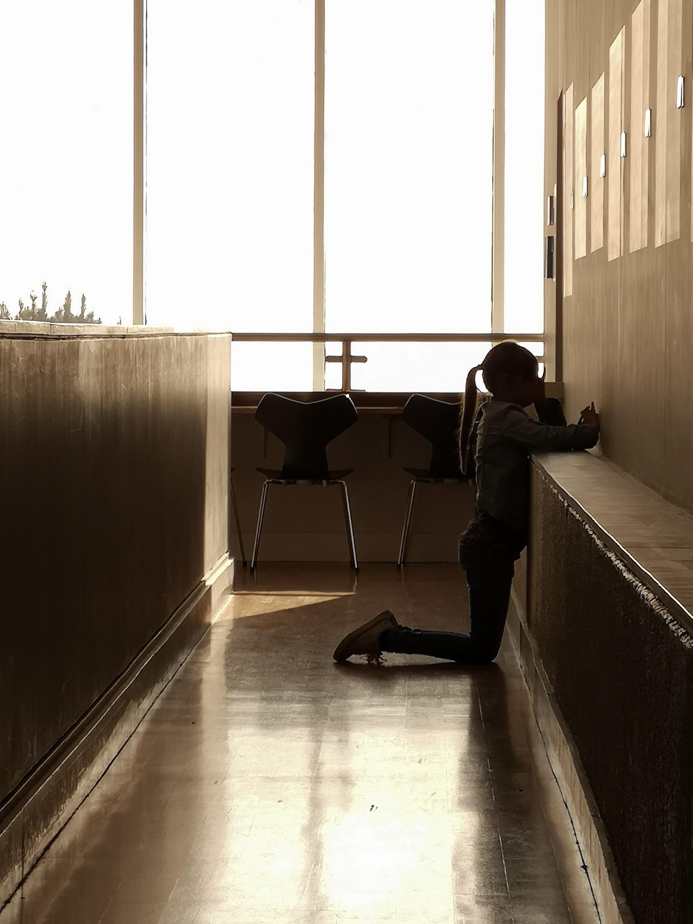 Silhouetted child kneeling at a hallway ledge playing on an iPhone, backlit by large windows; two empty chairs sit in front of the windows, and sunlight reflects on the polished floor. A safety barrier in front of the windows resembles a cross.