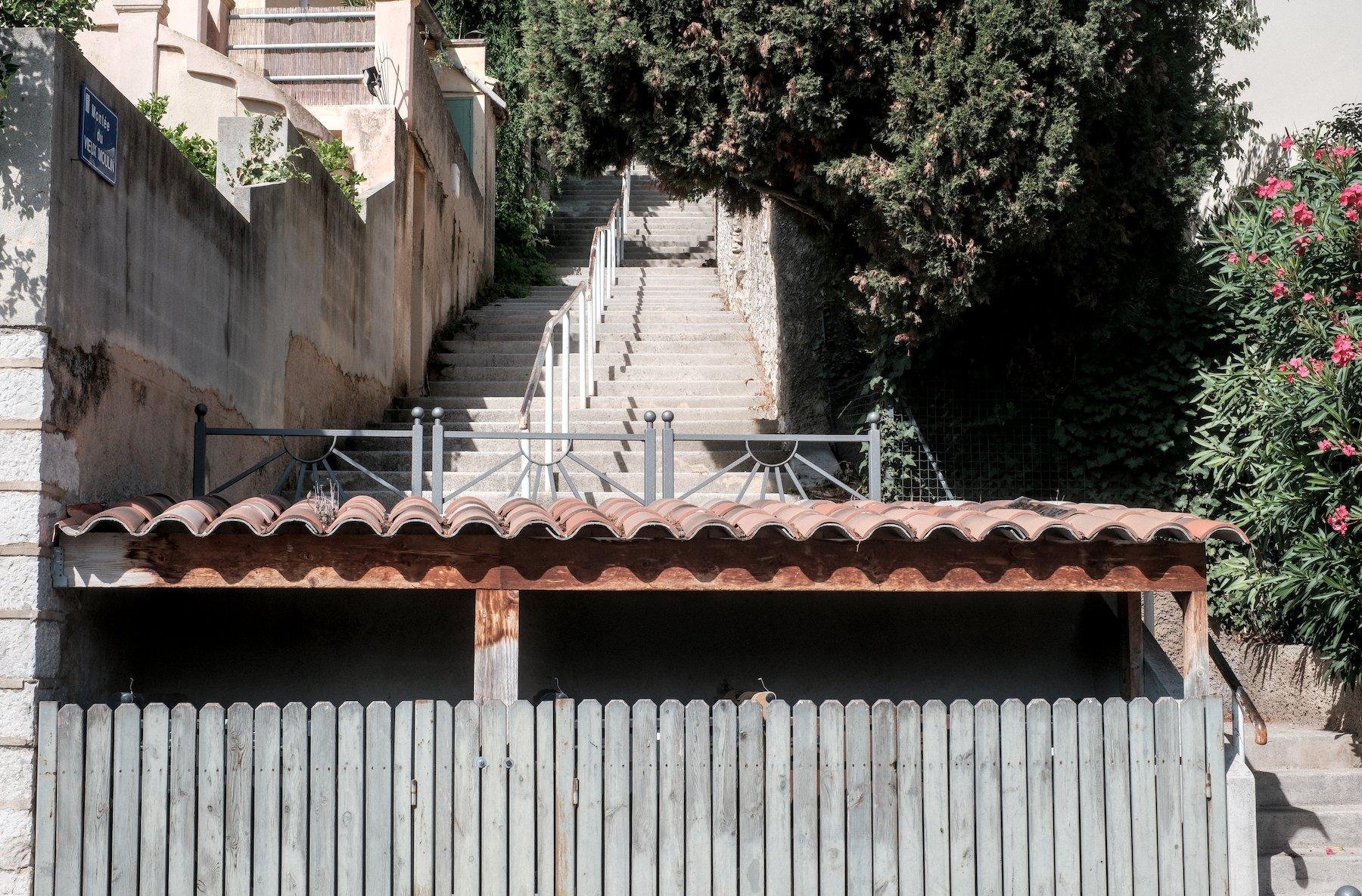 Photo horizontale. Un escalier en ville contourne un abris au toit en tuiles arrondies, et une palissade en bois gris, derrière laquelle on distingue des bouts de poignets de poubelles