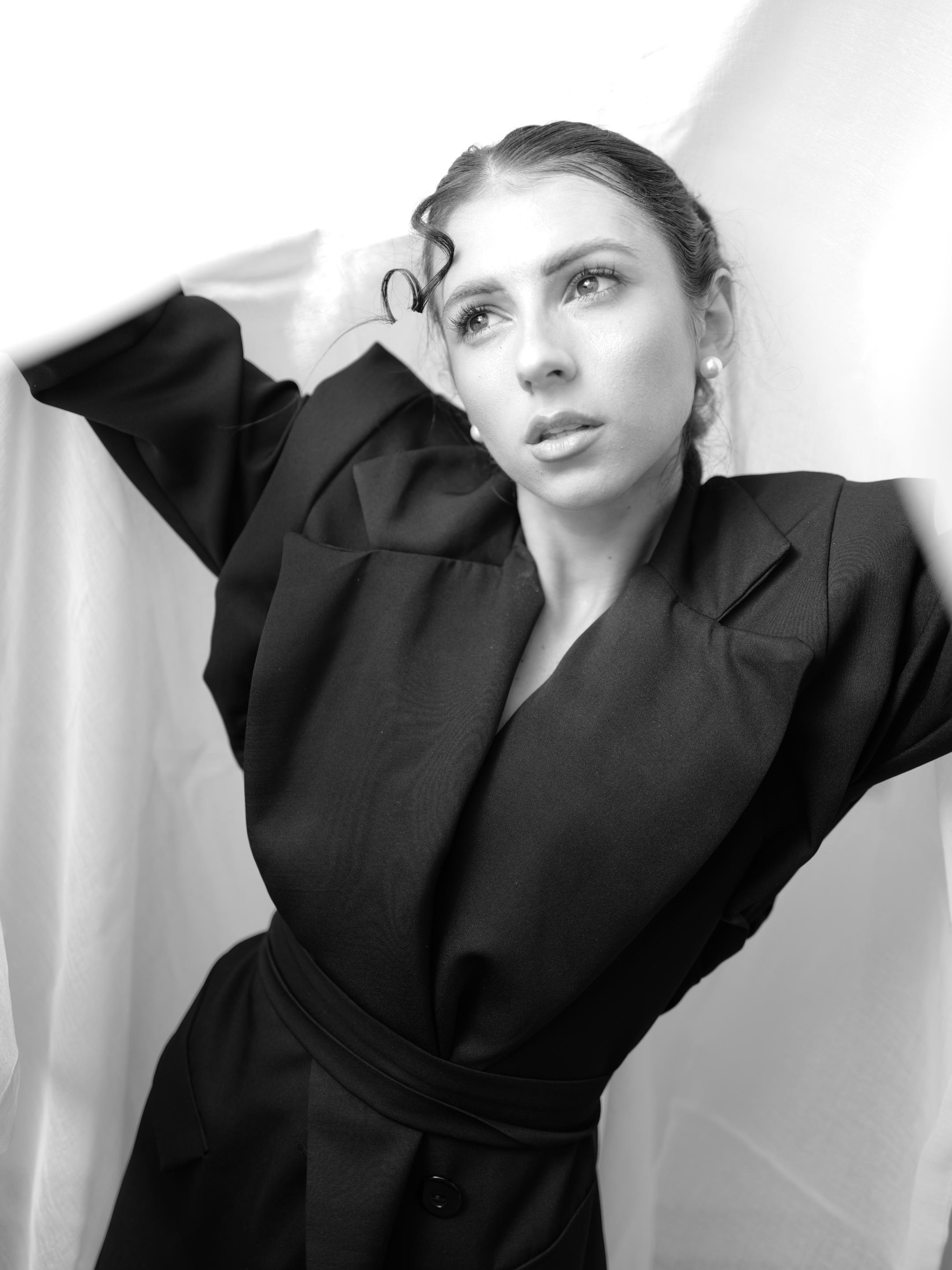 Black-and-white portrait of a woman in a belted dark blazer with pearl earrings, hair pulled back with a curled strand on her forehead, arms raised against a light fabric backdrop while looking off to the side.