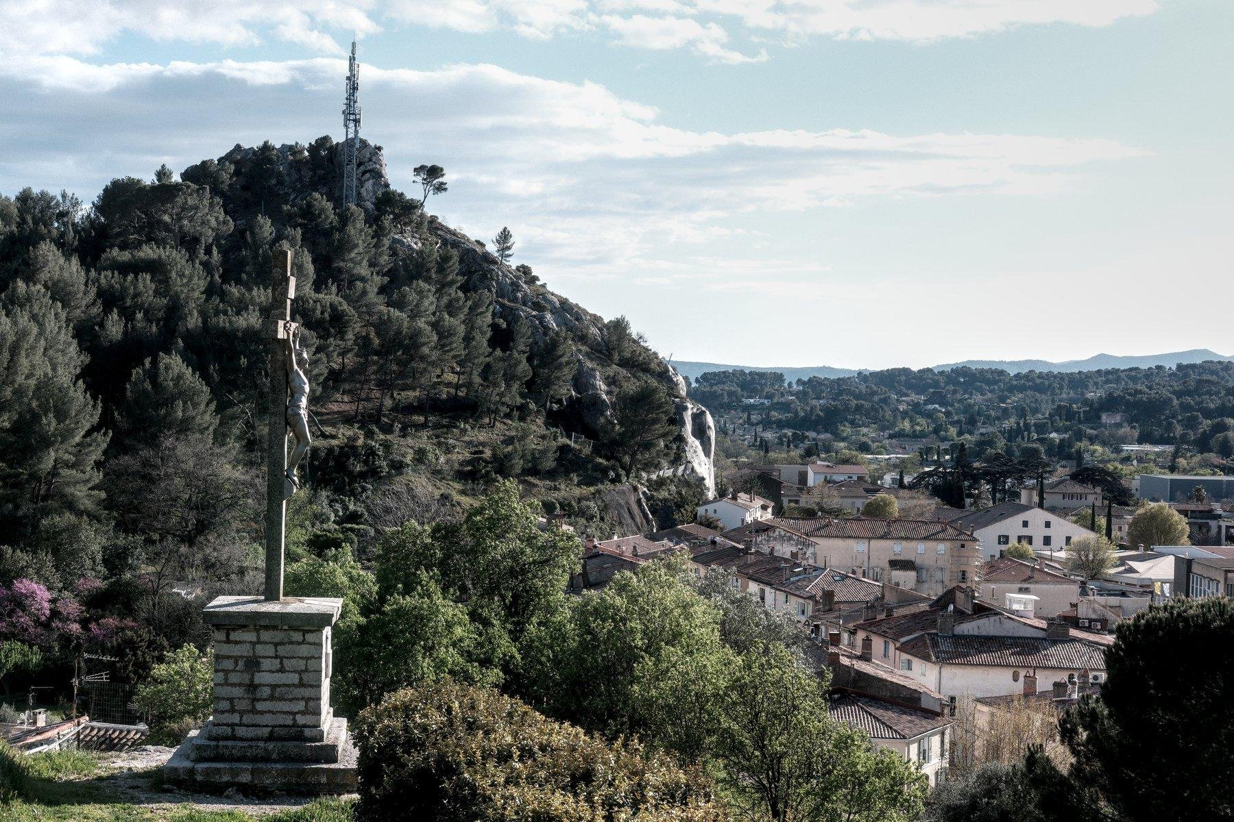 Photo horizontale. Sur la gauche, une croix avec la statue de Jésus crucifié. À droite, vu plongeante sur le village de Roquevaire. Au fond un bout du Garlaban, la montagne de la région, avec une antenne de communication dessus