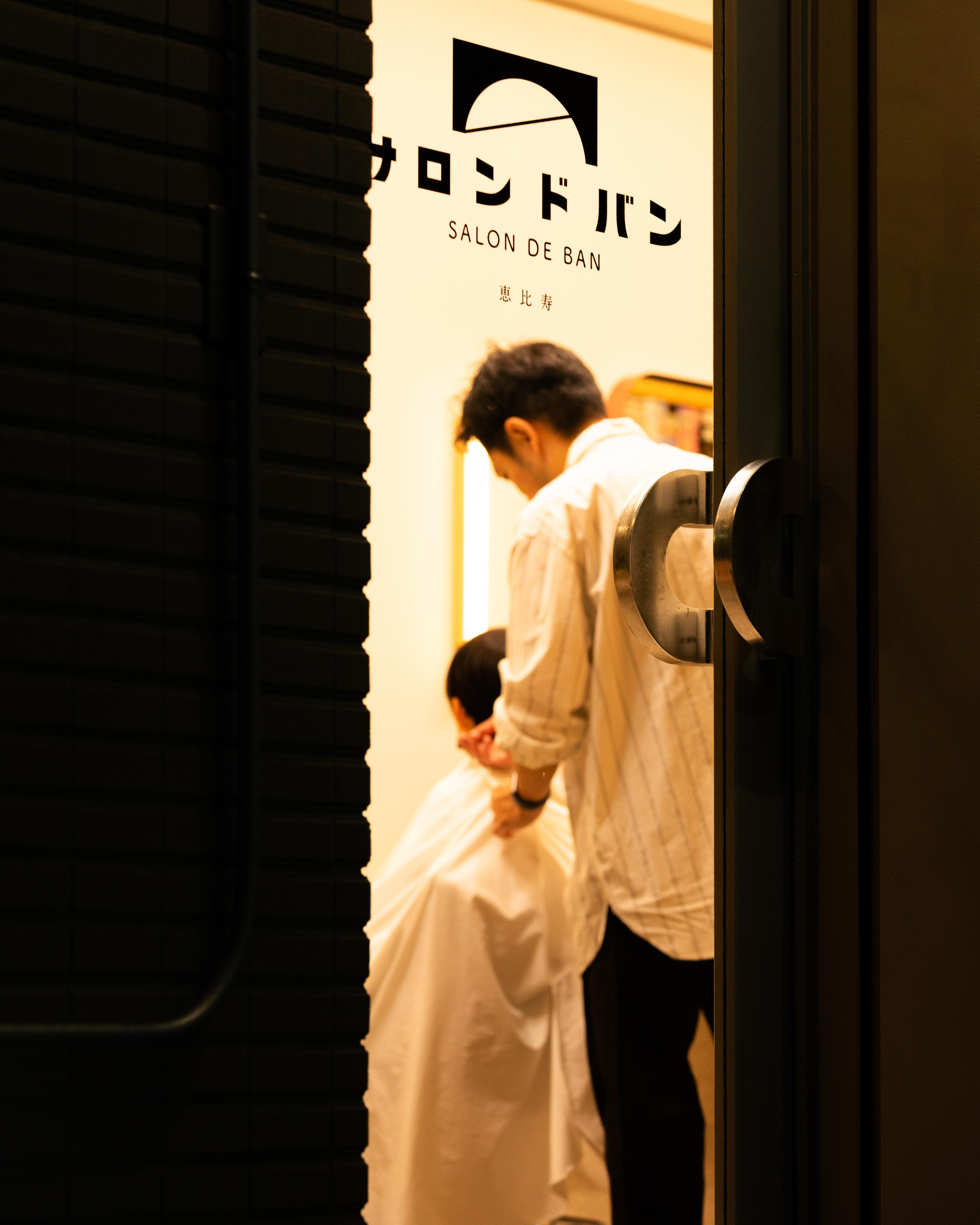 A barbershop interior seen through a partially open door, with a person in a striped shirt attending to a seated customer covered by a white salon cape. "Salon de Ban" is written on the wall, along with stylized Japanese text and a logo featuring a semicircle. Dim, warm lighting gives a cozy ambiance.