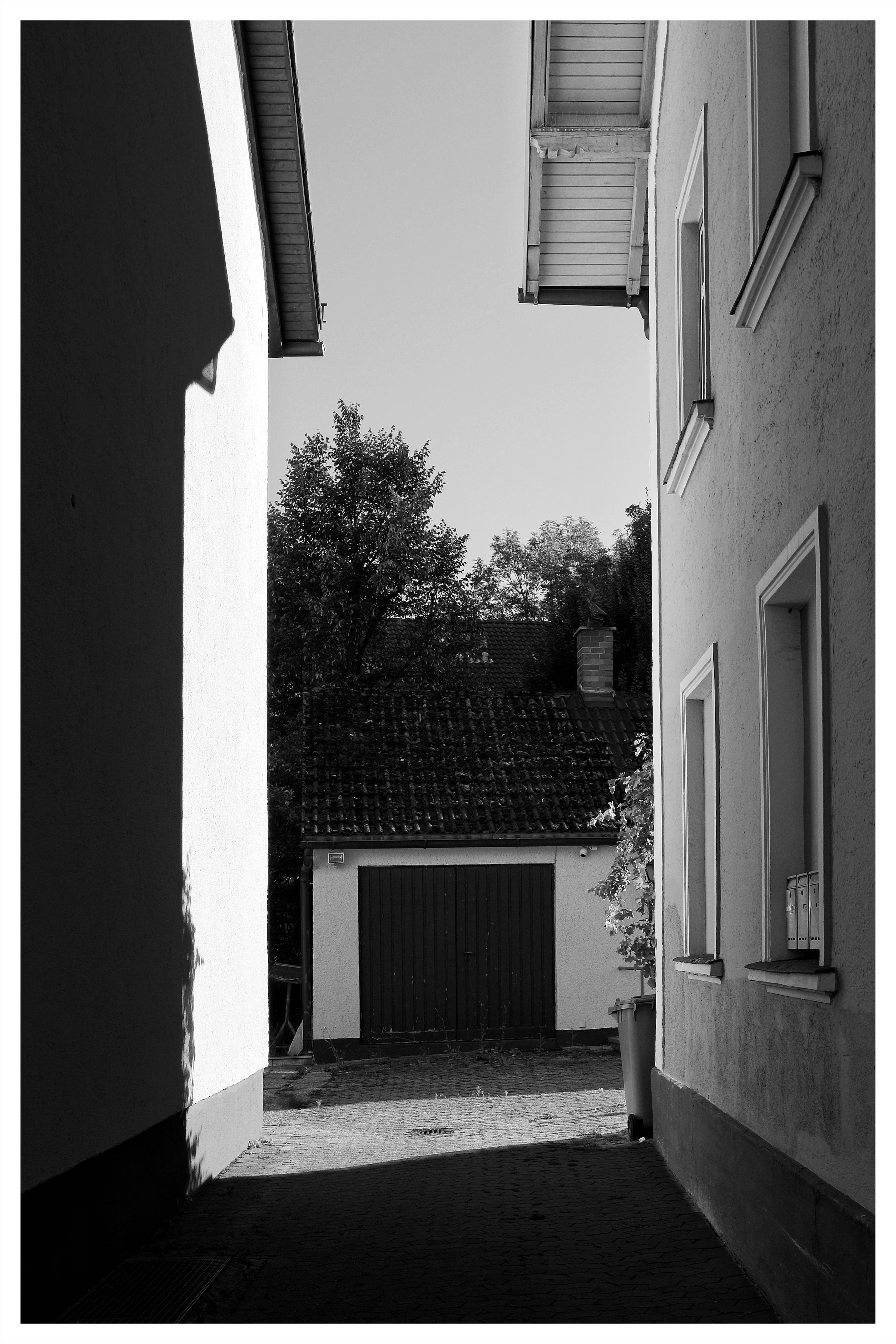 Black-and-white view down a narrow alley between two buildings, with windows and eaves on the right and a shadowed wall on the left; at the end is a small garage with double doors beneath a tiled roof, trees rising behind it, and a trash bin near the right edge.