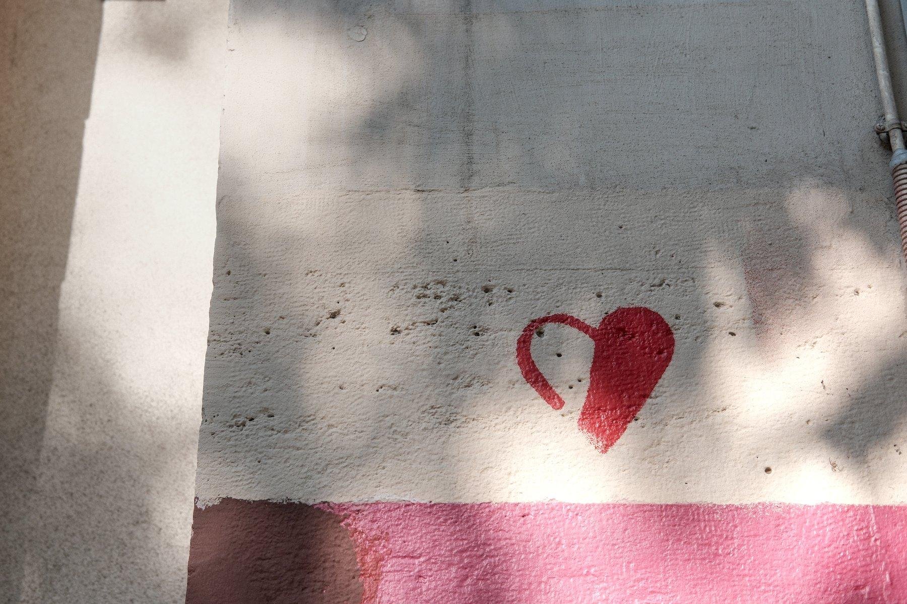 Photo horizontale. Un mur blanc, avec en bas de la photo une bande rouge, au dessus, un streetart peint d’un cœur à moitié plein, blanc et rouge.