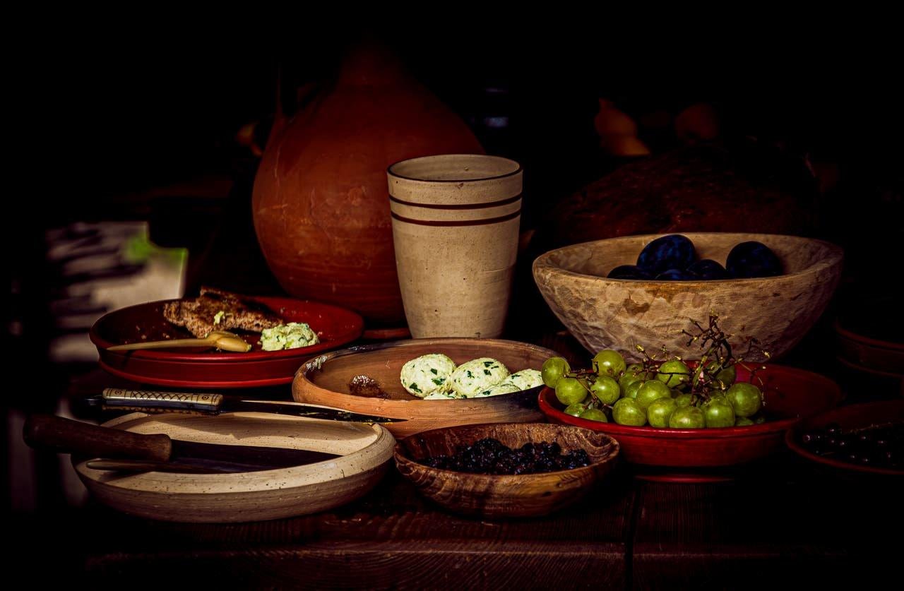 Still-life arrangement on a dark wooden table with a clay jug and a tall ceramic cup in the center, surrounded by bowls and plates holding green grapes, dark berries, blue plums, cheese balls, bread, and wooden utensils against a dark background.