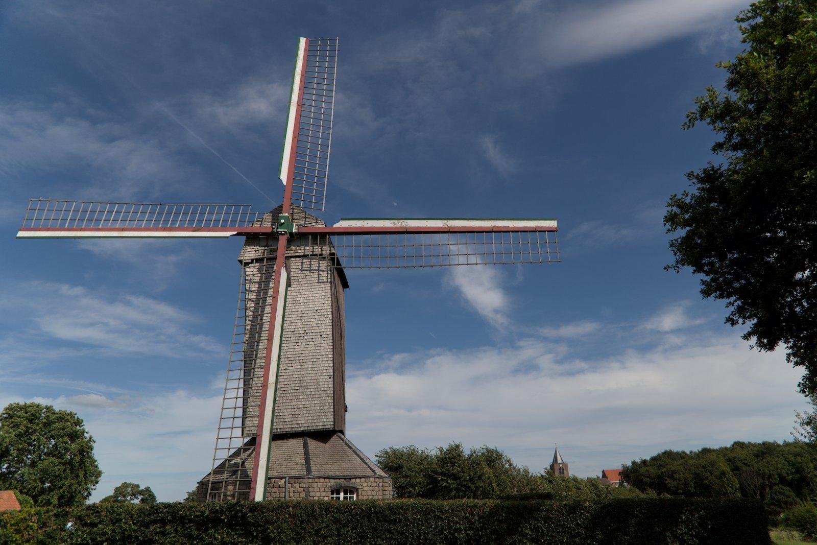 Photo format paysage montrant un moulin à vent entier, de face avec un ciel bleu derrière. Le bas de l’image montre qu’il se trouve pas loin de forêt et une haie bien taillée ferme l’image en bas