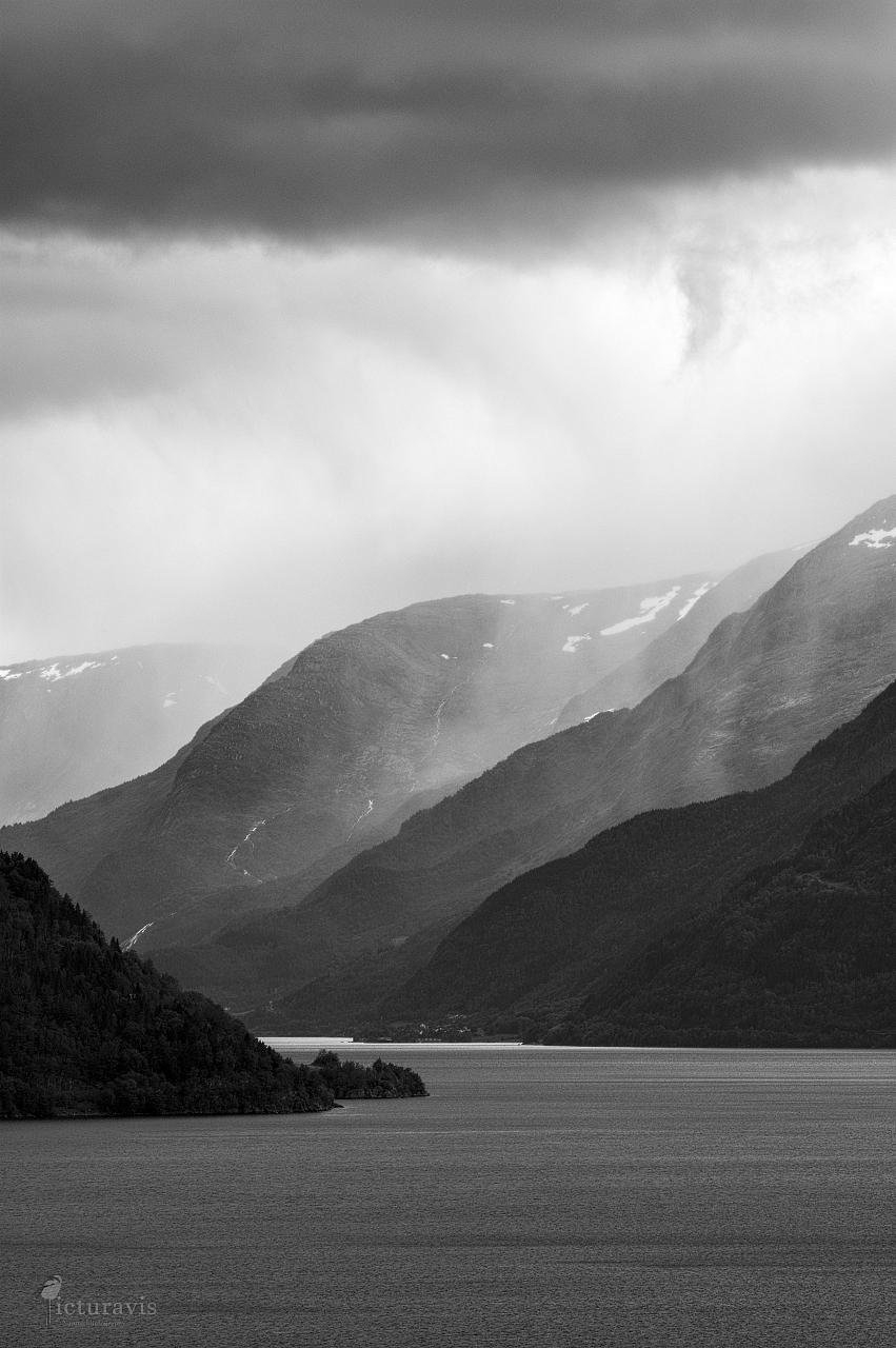 A vertical grayscale image of a fjord, where the sea cuts deep into the landscape between mountains. Clouds hang over the mountains, raining onto the landscape below, creating a dramatic image.