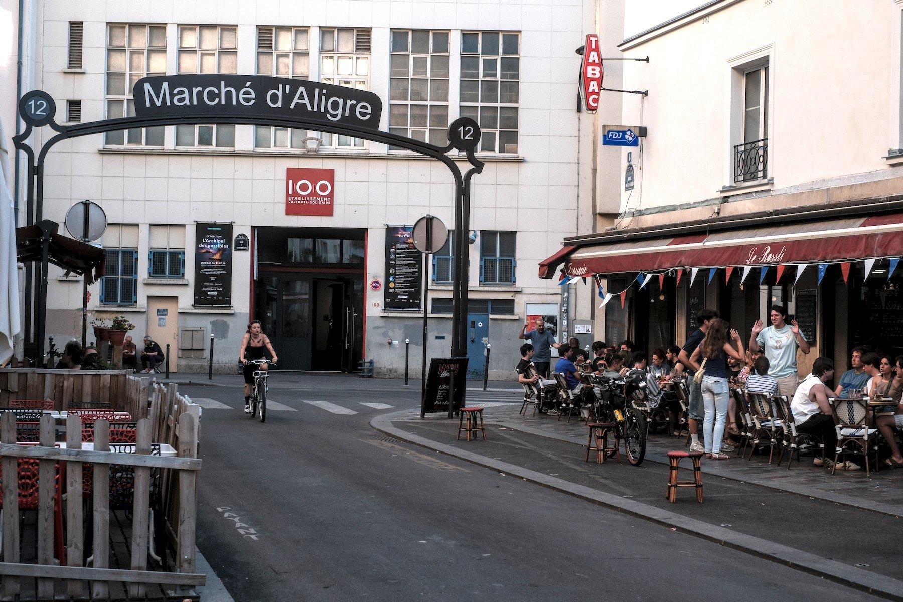 Photo horizontale. Une rue dont l’entrée est surmonté d’une grande arche verte sombre faisant office de porche, avec au sommet un panneau marqué « Marché d’Aligre ». Une cycliste passe en dessous, on voit des terrasses des deux côté de la rue