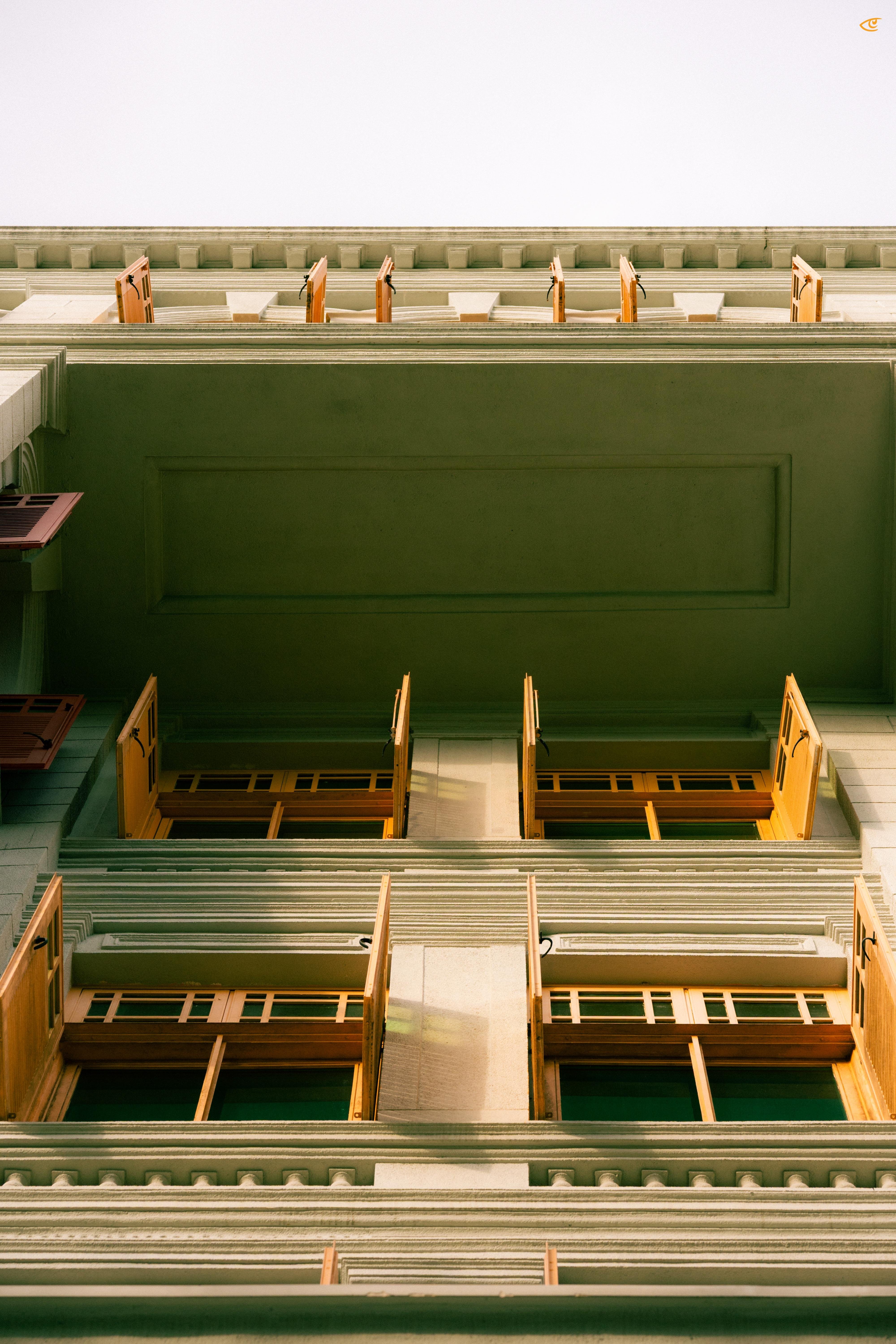 Upward view of a multi-story building facade with pale walls, decorative cornices, and rows of windows with wooden shutters opened outward, framed against a bright sky.