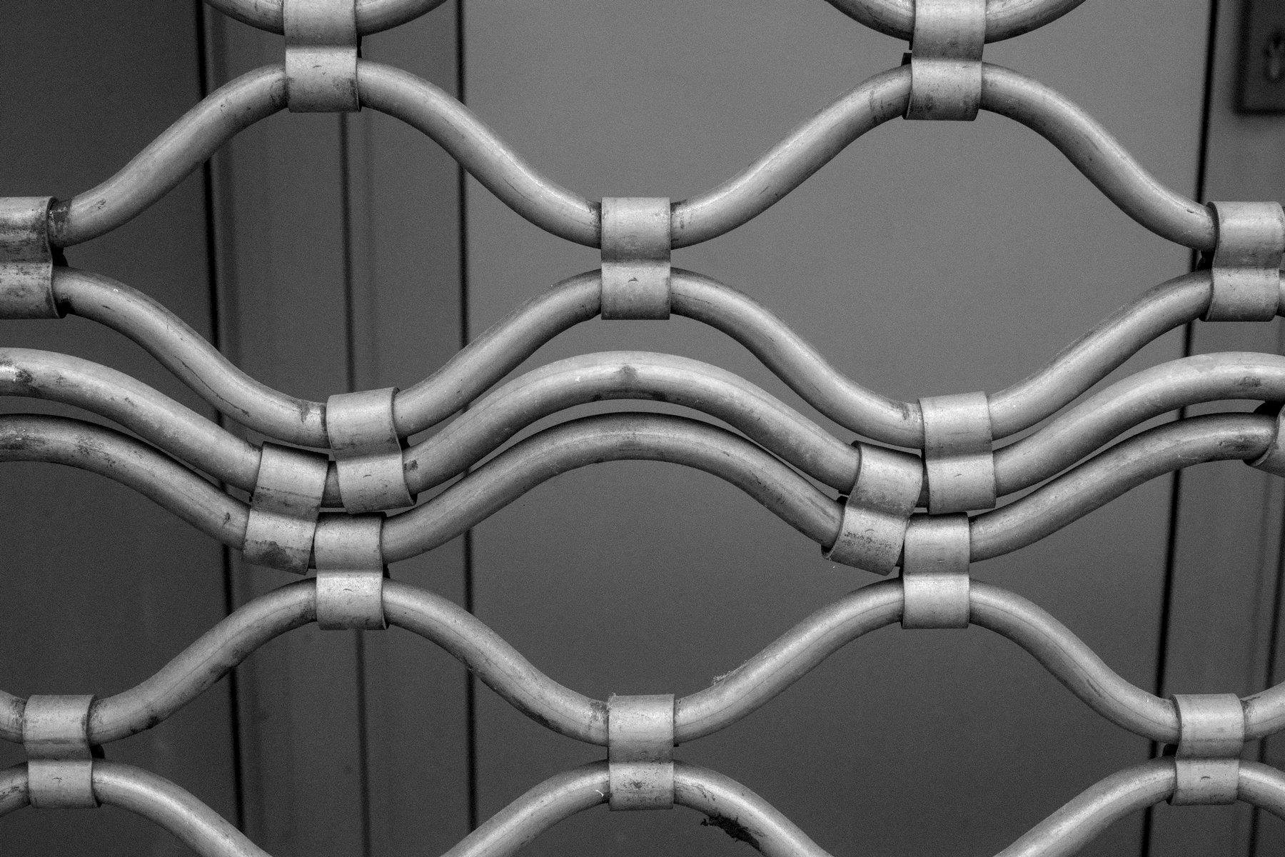 Photo horizontale en noir et blanc. Gros plan sur quelques mailles d’une grille rideau de sécurité, avec des mailles qui sont comme compressées