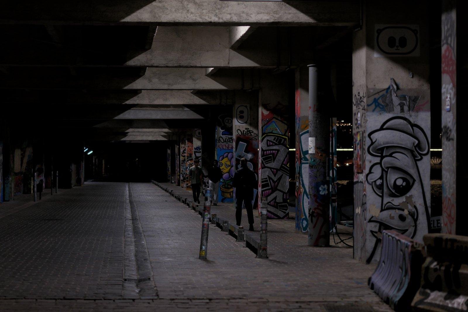 Photo horizontale, de nuit. Un rue piétonne couverte, dont les piliers tenant le toit sont couvert de street art