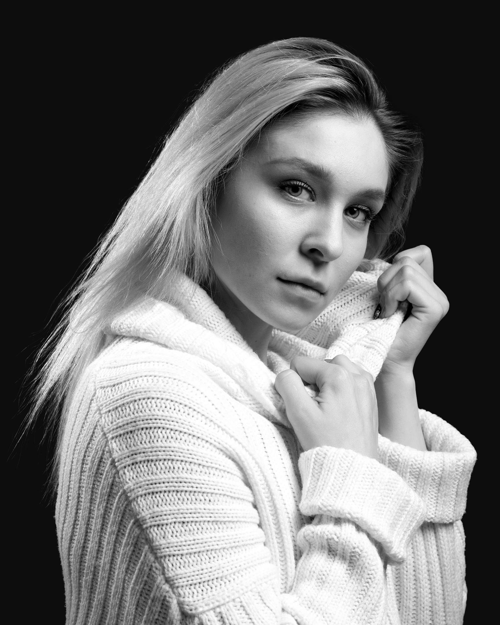 Young woman with long, light hair, wearing a thick, textured knit sweater. She is gently pulling the lapel of the sweater with both hands, gazing directly at the camera in a poised manner. The background is plain and dark, which contrasts with her light hair and sweater. The image is in black and white, emphasizing the texture and shadows.