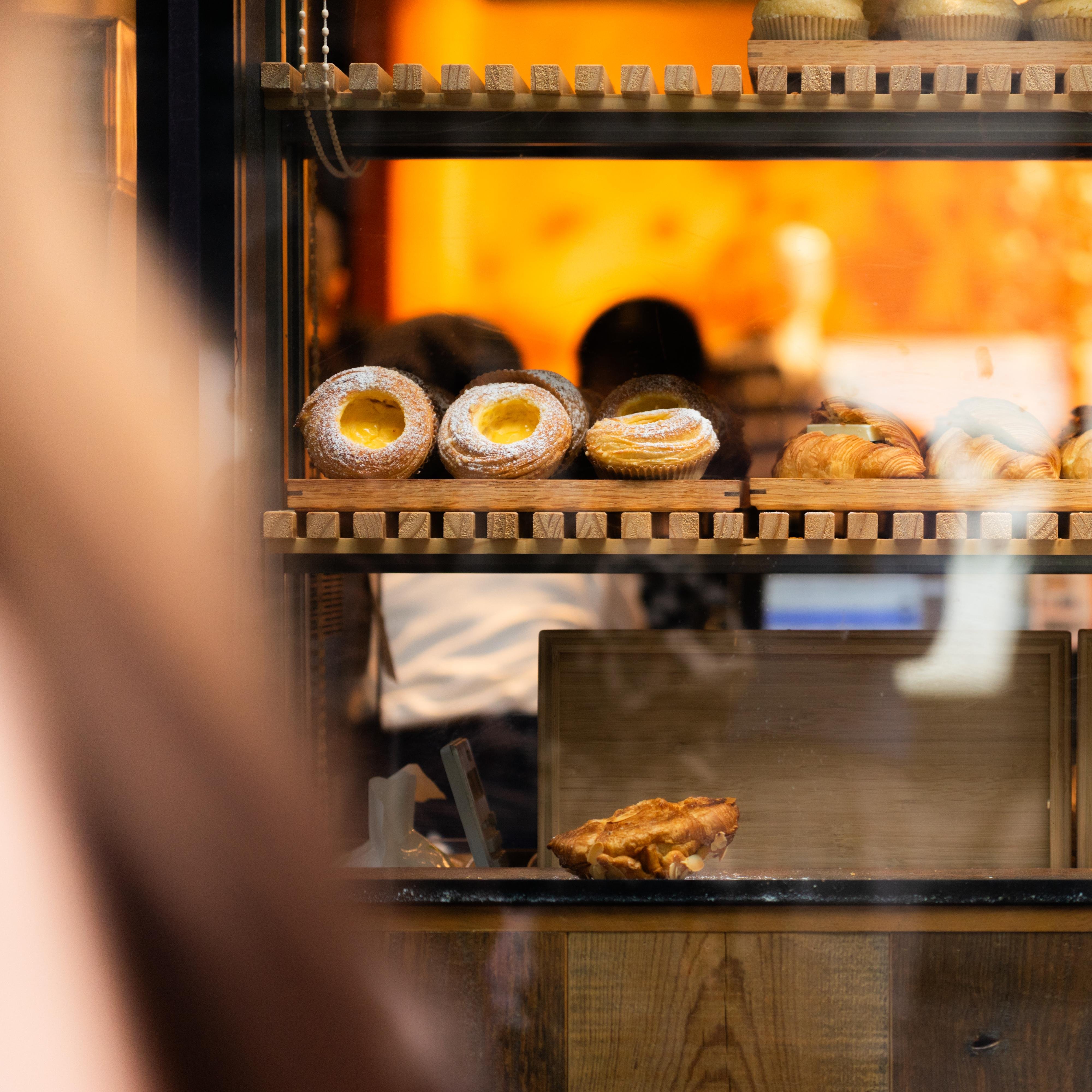 Bakery display with a wooden shelf holding pastries, including ring-shaped pastries with yellow centers sprinkled with powdered sugar, small tarts, and croissants. The background features an orange glow and silhouettes of people behind glass.