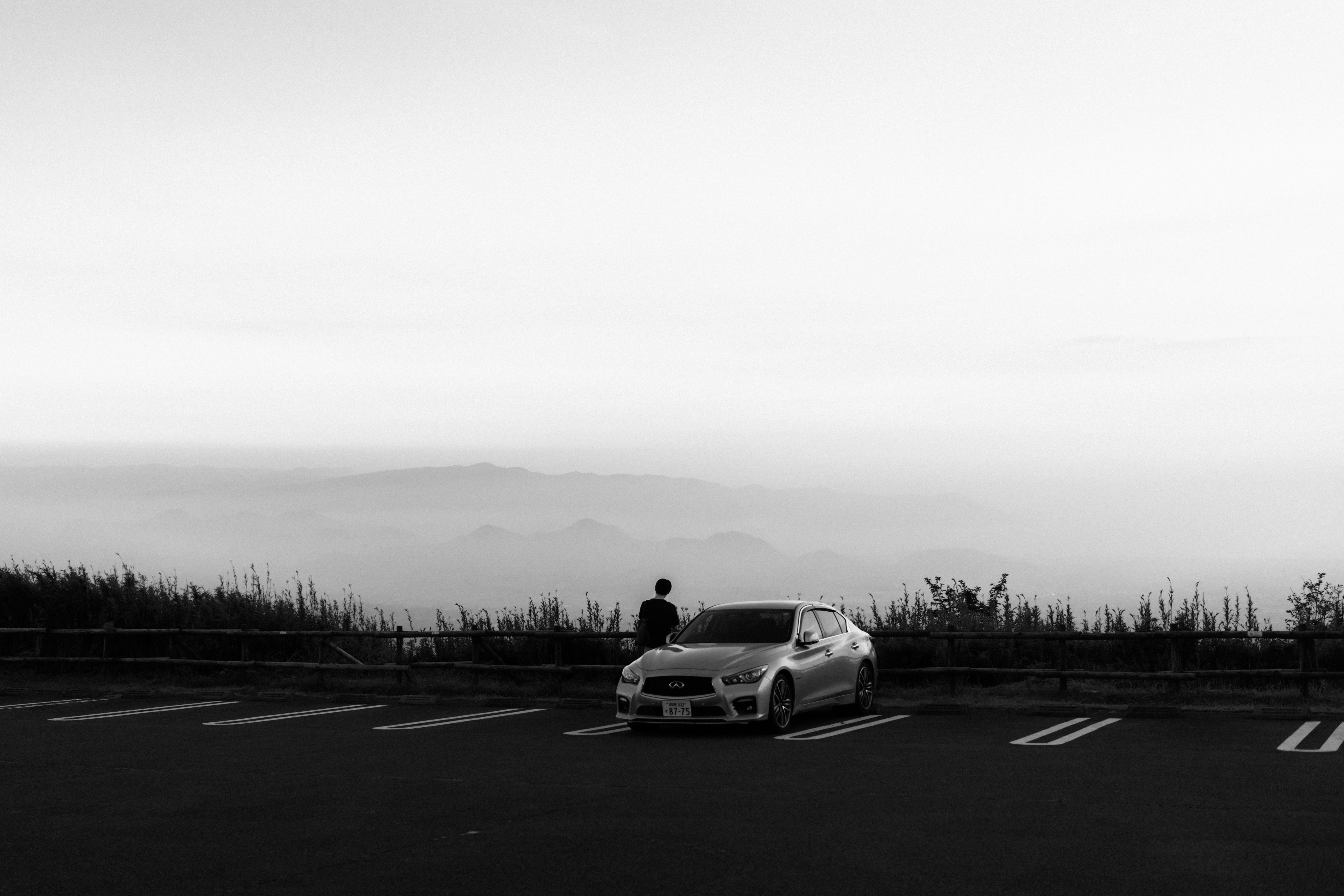 Black and white photo of a lone car parked in an empty lot, with a person standing next to it. Beyond the parking lot, a railing separates the area from a misty landscape, featuring distant, hazy mountains under a clear sky.