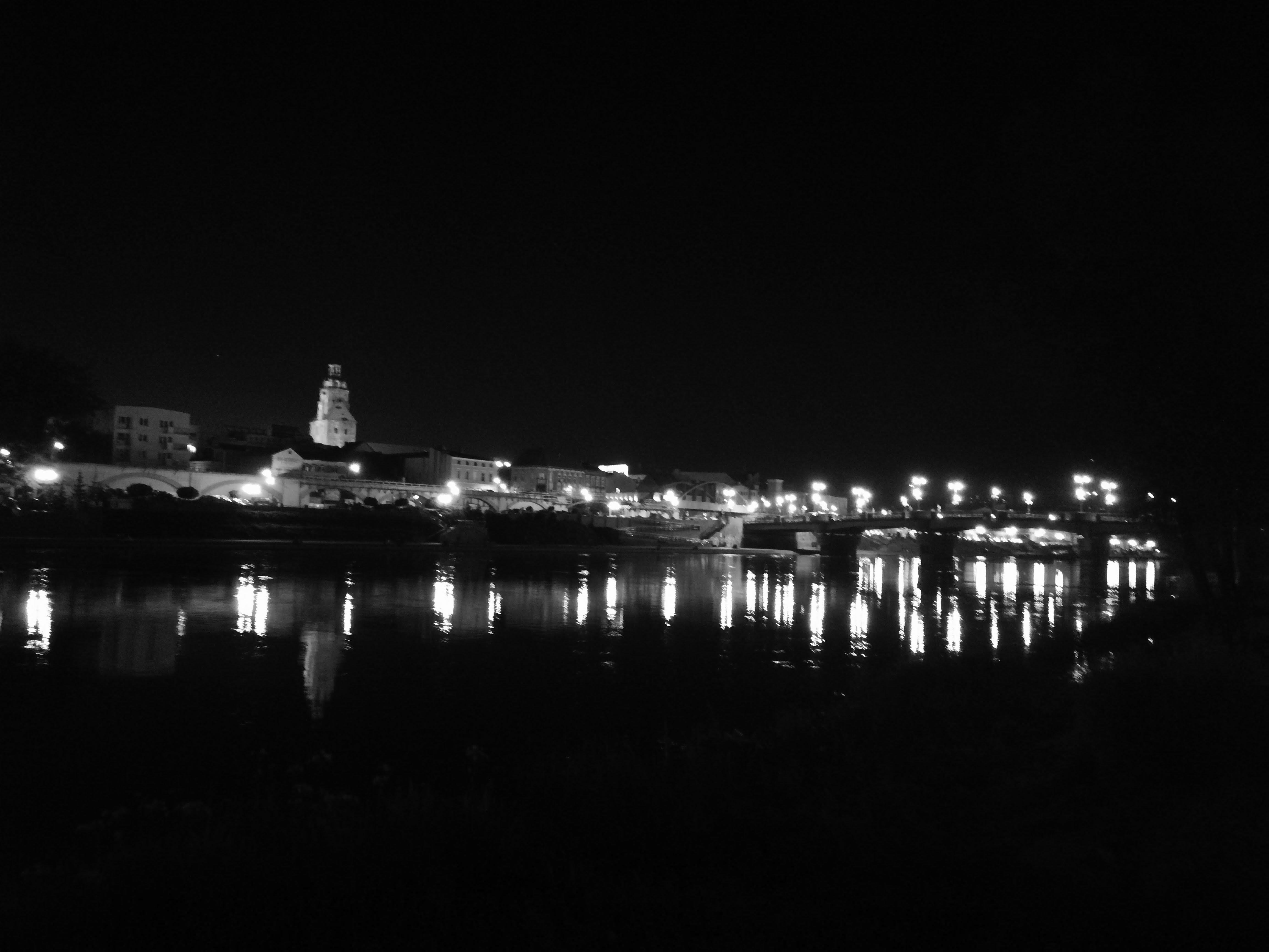 The image depicts a nighttime cityscape with buildings and structures illuminated by streetlights. There is a large river or body of water in the foreground, reflecting the lights from the buildings and streetlights. A bridge spans the river, also lit up by streetlights. The overall setting suggests an urban area, possibly a historic town. The image is in black and white.