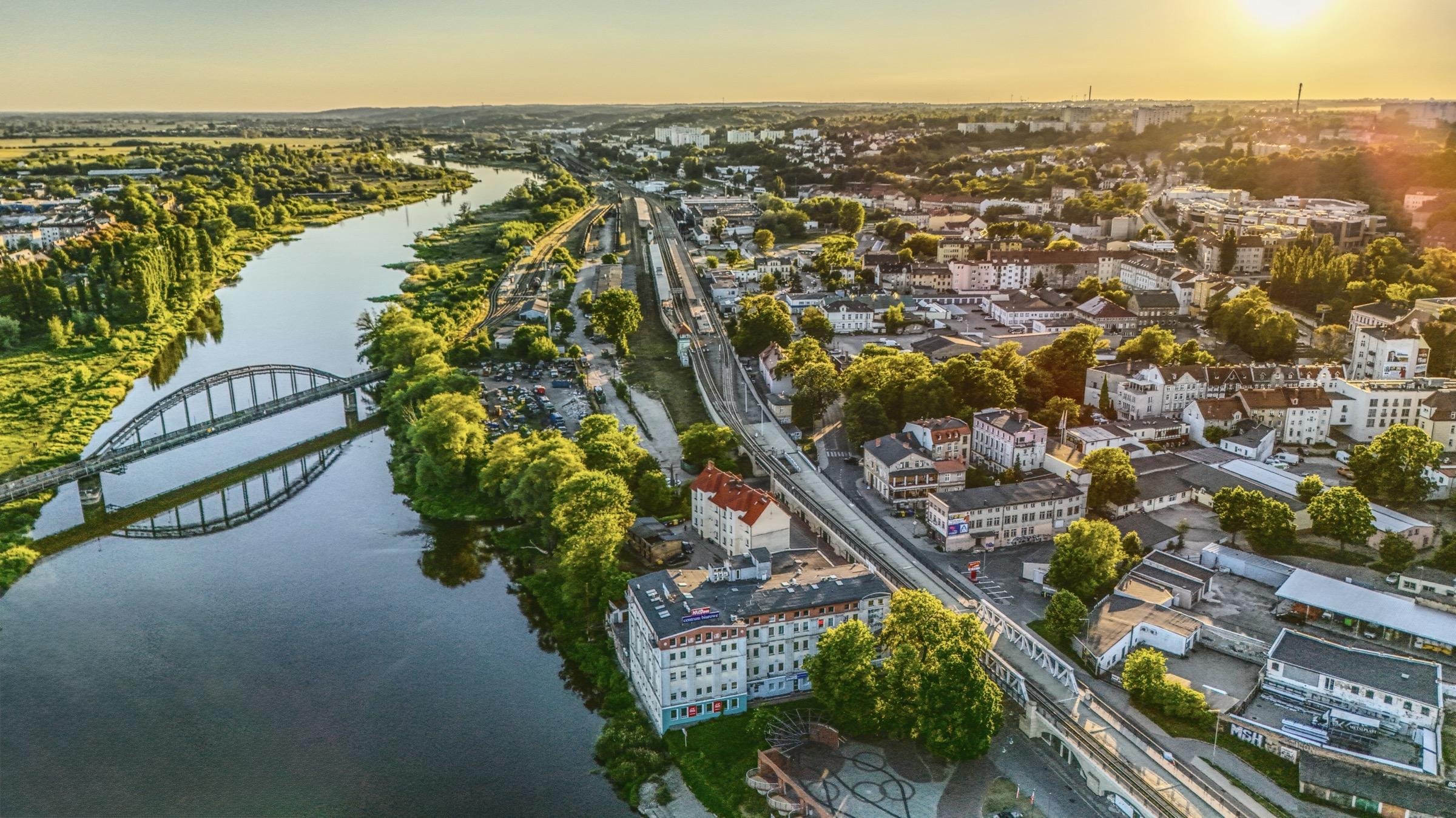Aerial view of a cityscape at sunset, featuring a winding river on the left bordered by lush green trees. A prominent bridge spans the river, leading to a developed urban area with numerous buildings and streets. Train tracks run parallel to the river. The sun sets in the background, casting a warm golden hue over the scene.