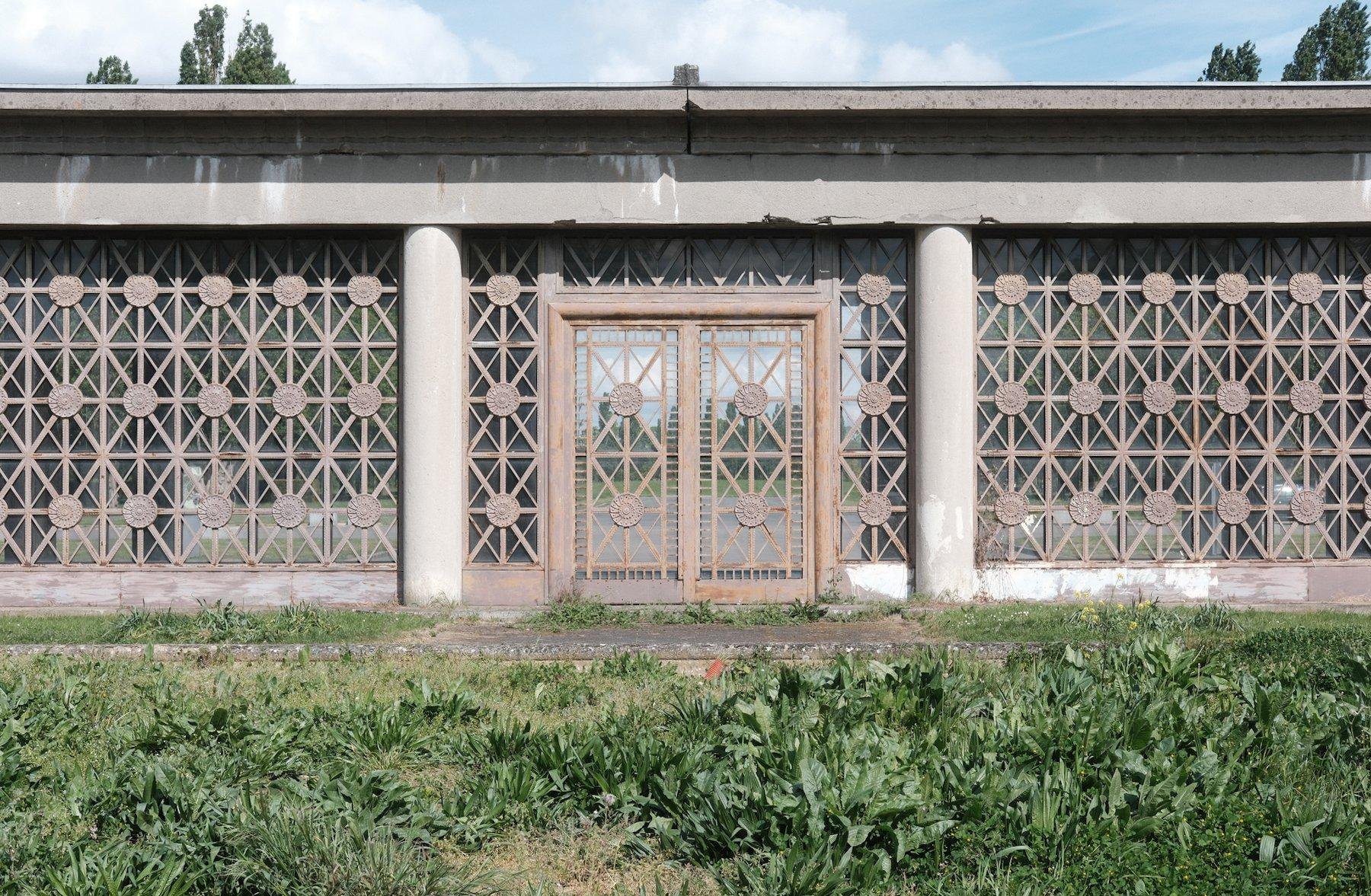 Photo horizontale. Une porte dans une barrière massive faite d’un maillage en losange avec des nœuds marqués d’un disque, le tout de couleur rouille clair, maillage dans une structure en béton gris, avec 2 poteaux cylindriques formant un linteau à la porte
