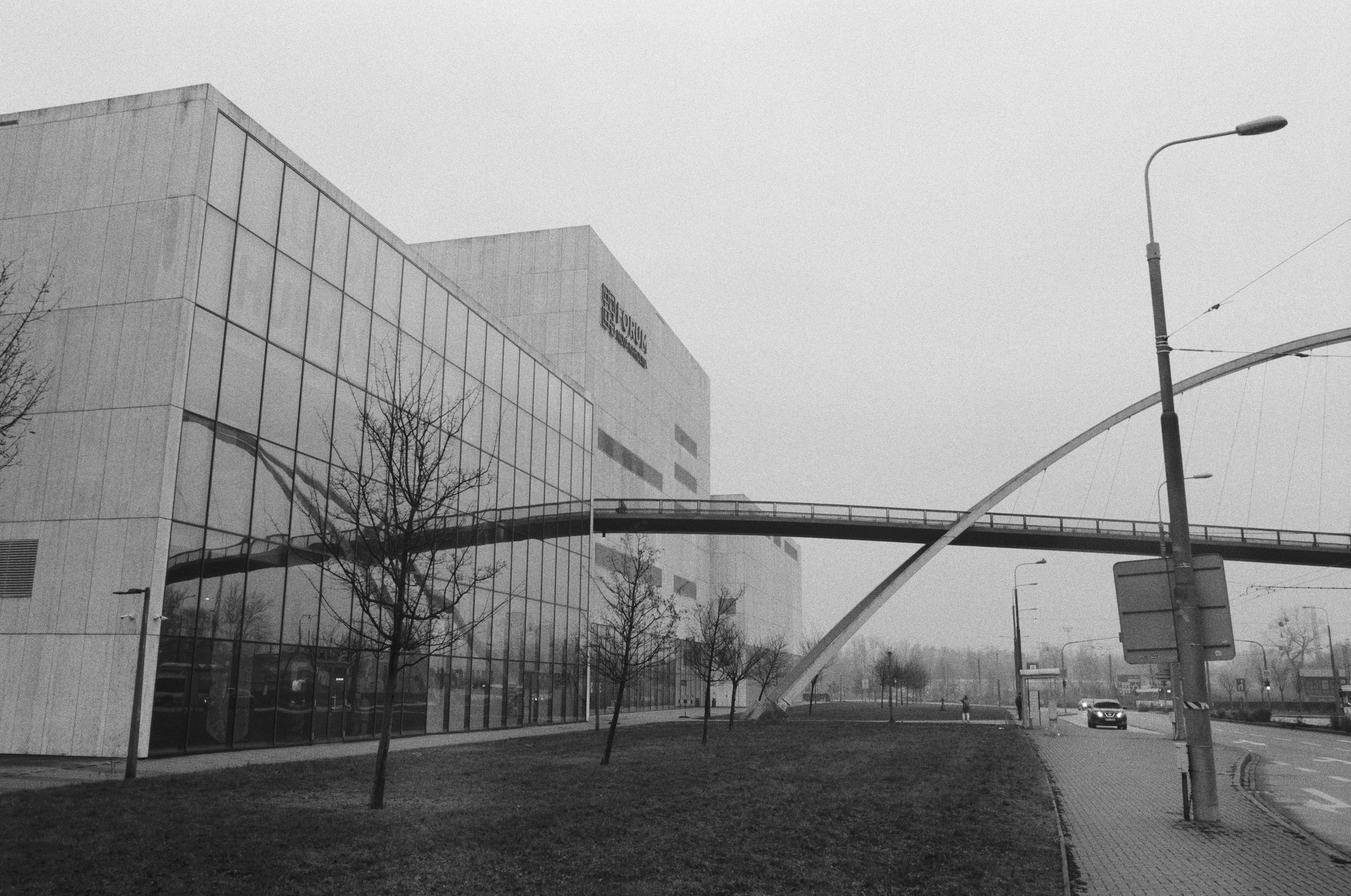A monochrome photograph of a concrete building with a full glass side and a pedestrian bridge leading towards the building reflecting until the glass side