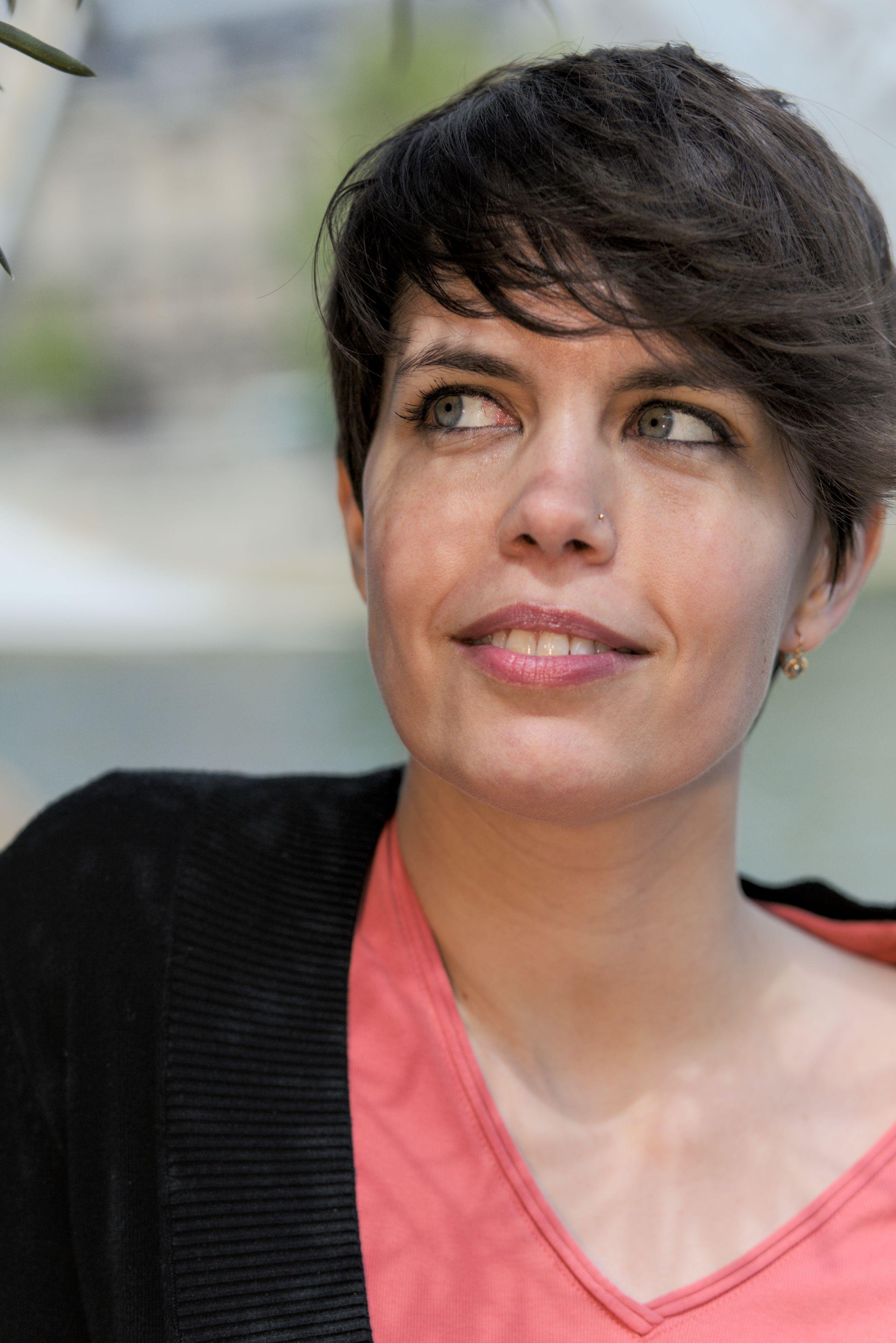 Photo verticale. Plan serré sur le visage d’une jeune femme brune qui porte un haut rouge et un gilet noir par dessus. Elle a son visage face à nous mais regarde une branche du coin des yeux vers la gauche de la photo