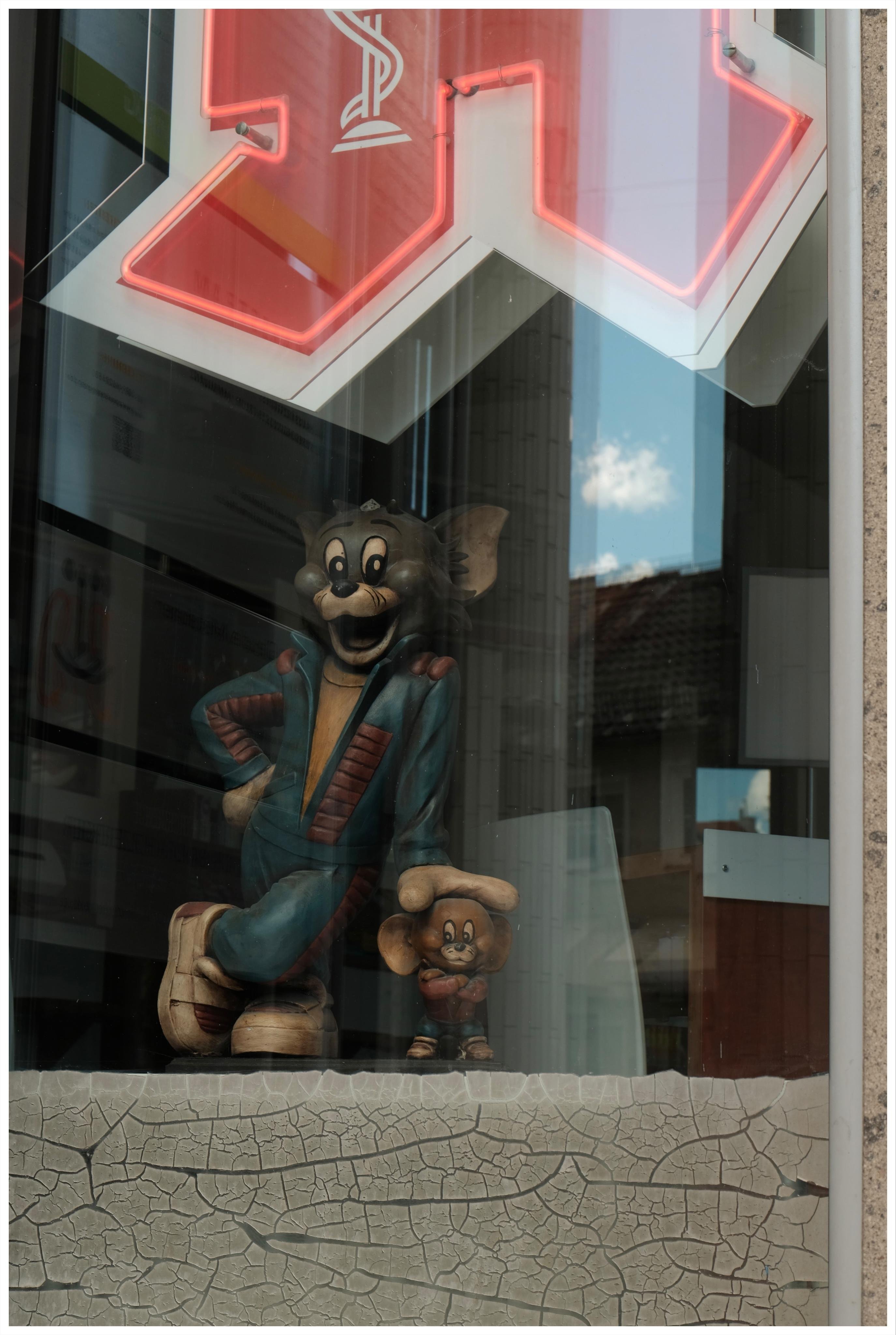 Shop window of a pharmacy displaying a figure of Tom and Jerry where Tom put a hand on Jerry and casually leans on him.
At the top of the shop window, the lower part of the rate pharmacy sign is visible. The lower part a cracked and torn paper seems to be glued to the shop window as a base layer for the shop display.