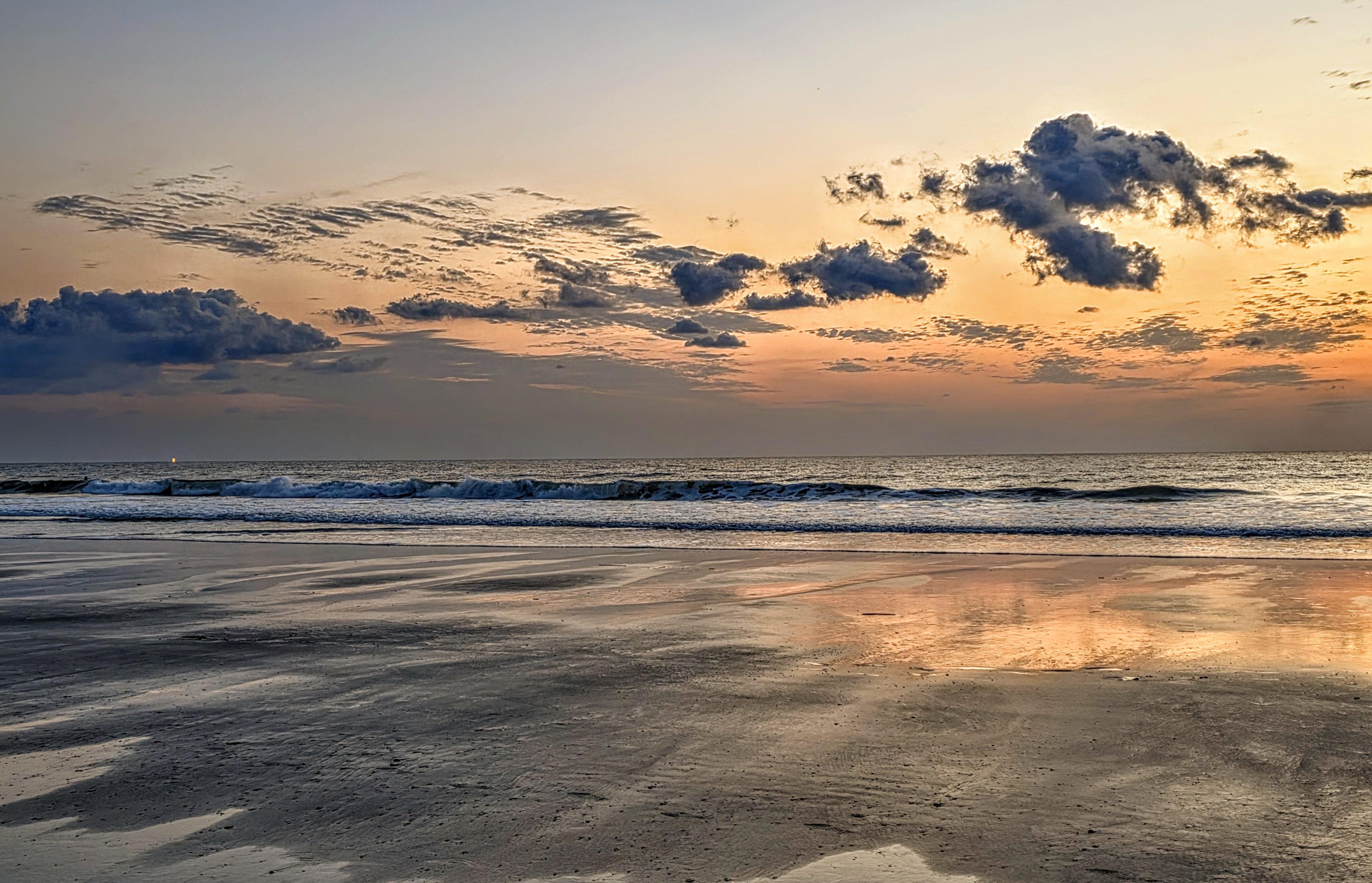 Sunrise over a tranquil beach with gentle waves approaching the shore. The sky is a gradient of orange to light blue, scattered with patches of clouds reflecting the sky's hues. Wet sand in the foreground mirrors the sky's colors, enhancing the serene atmosphere.